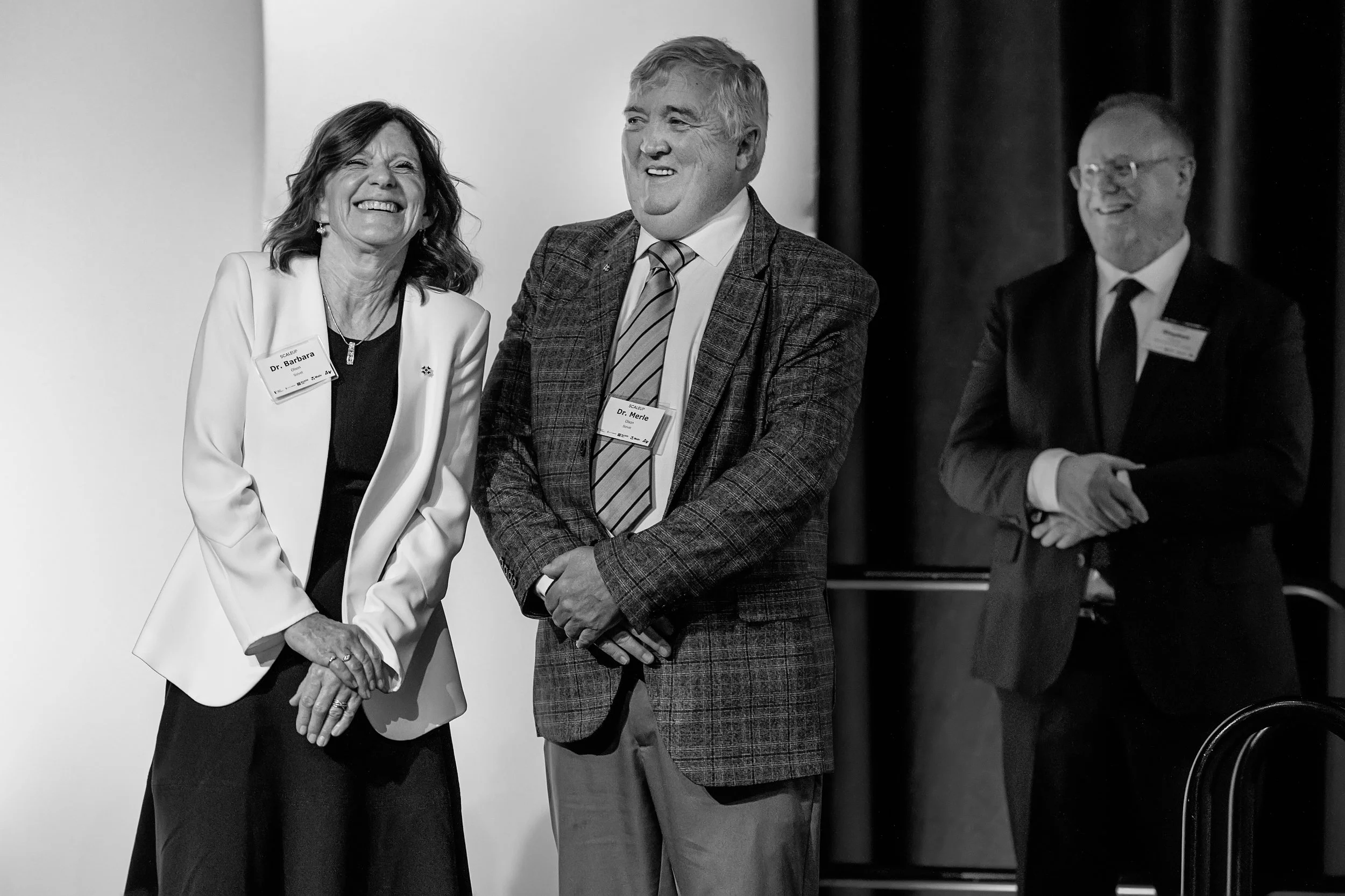 Three people standing together at a professional event, smiling and engaged in conversation. The woman on the left is wearing a blazer and a badge that reads 'Dr. Barbara,' the man in the middle is wearing a plaid blazer and a badge that reads 'Dr. Merle,' and the man on the right is dressed in a suit and glasses, smiling with his arms crossed.