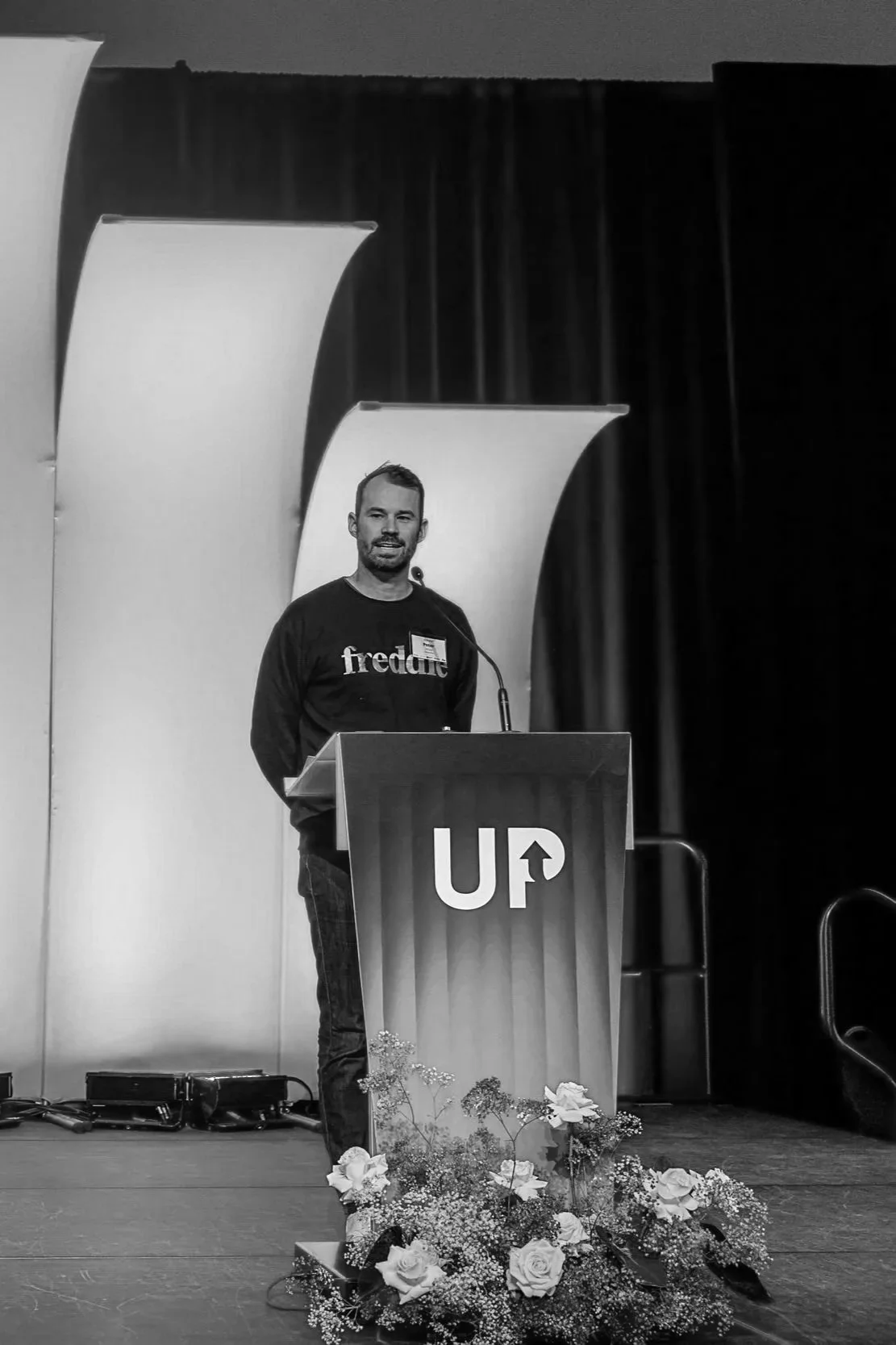 A man standing at a podium with a microphone on a stage, wearing a black sweatshirt with the word 'freddit' on it. The stage has abstract background panels and a floral arrangement in front of the podium.