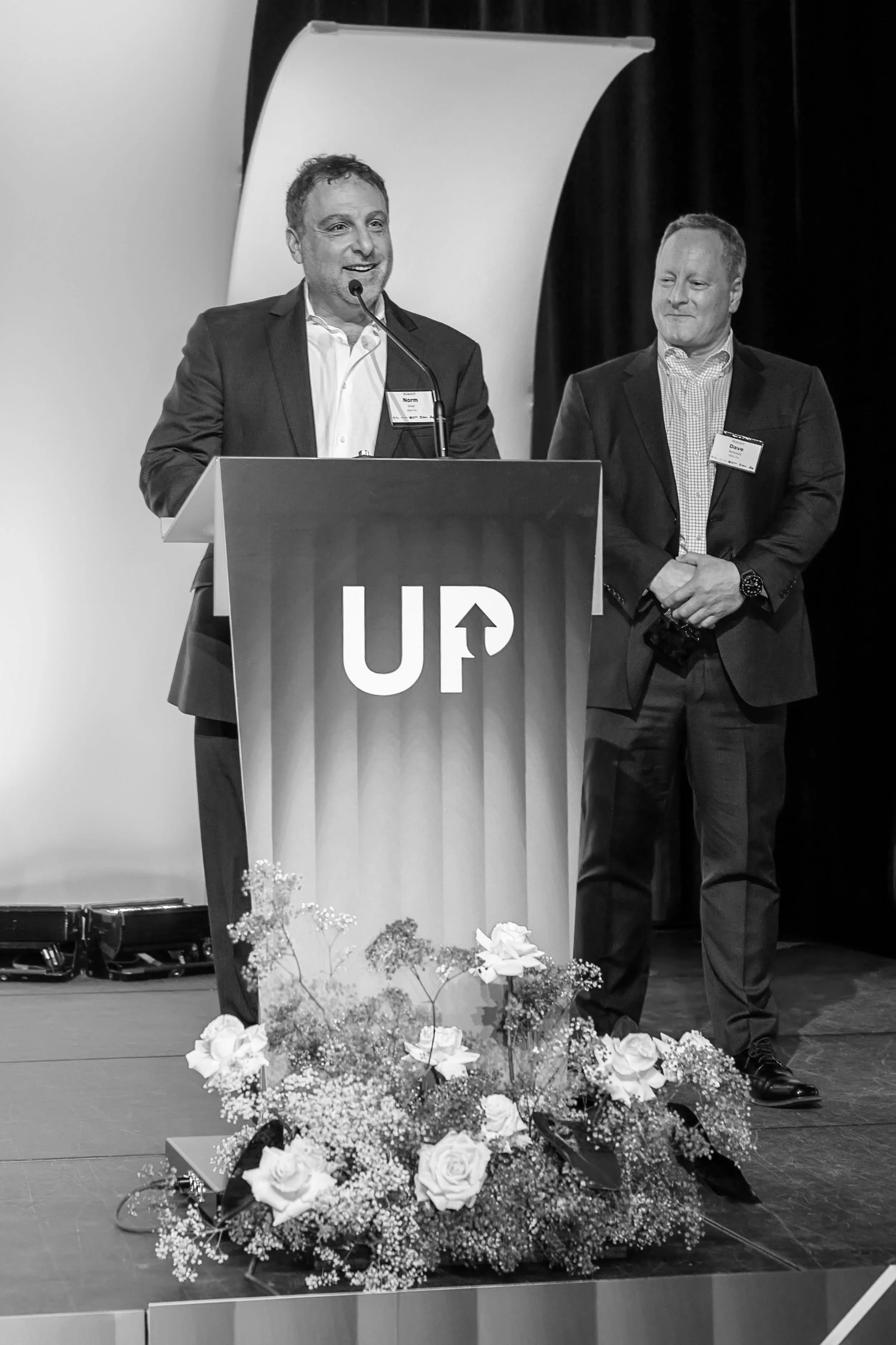 Two men in suits on a stage, one speaking at a podium with a microphone, the other standing beside, with a floral arrangement at the front of the stage. The podium has the word 'UP' with an upward arrow.