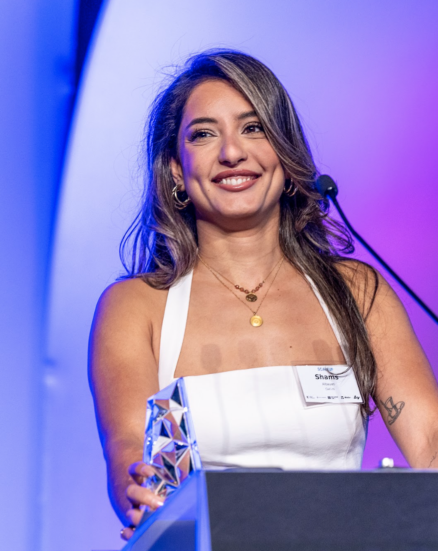 Owl.co award winner holding an award, wearing a white dress and layered gold necklaces, standing at a podium with a microphone in front of a purple and blue background.