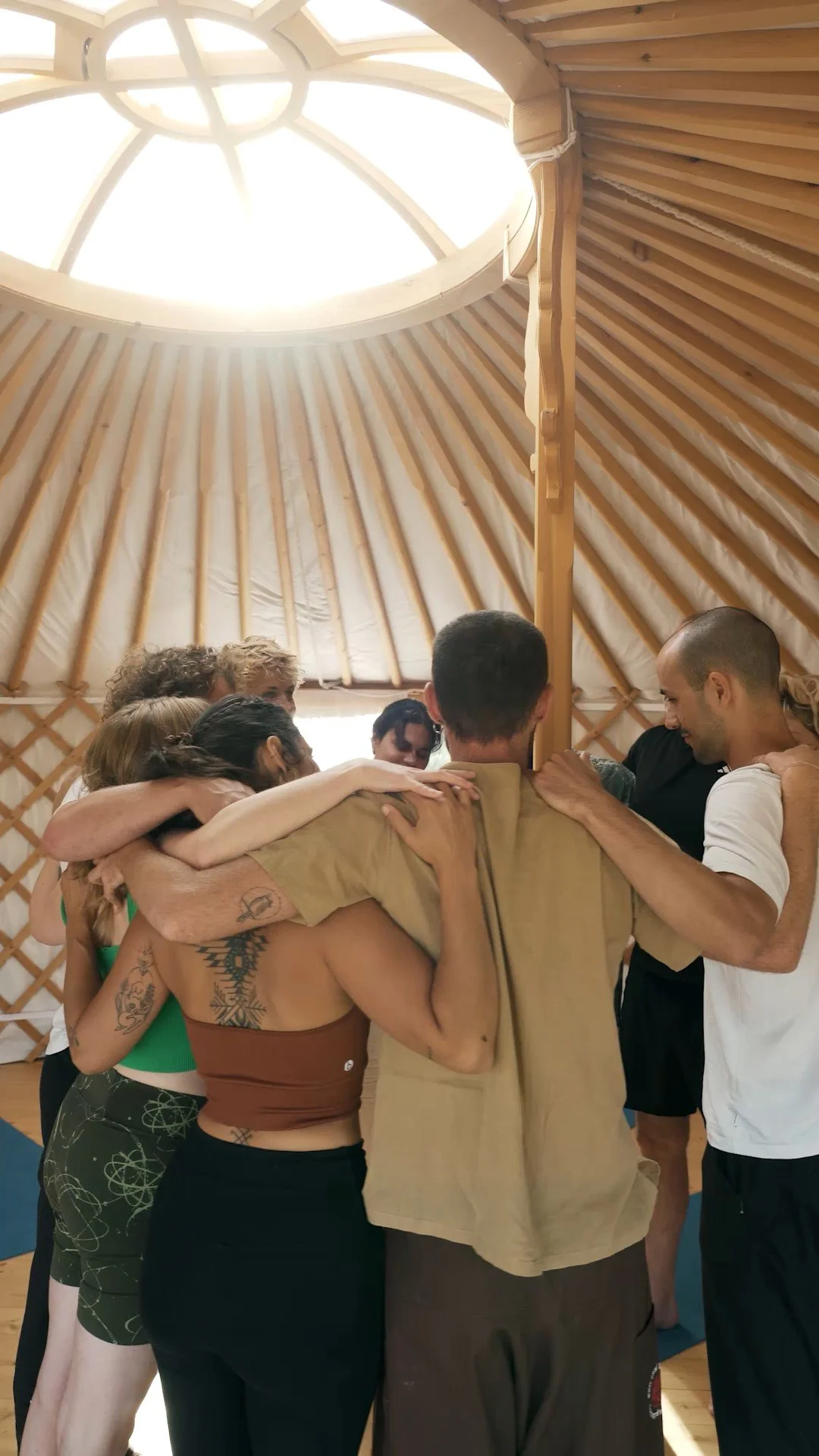 A group of people huddled together in a circle inside a Roundhouse with a high, domed ceiling, embracing each other, symbolizing unity or team spirit.