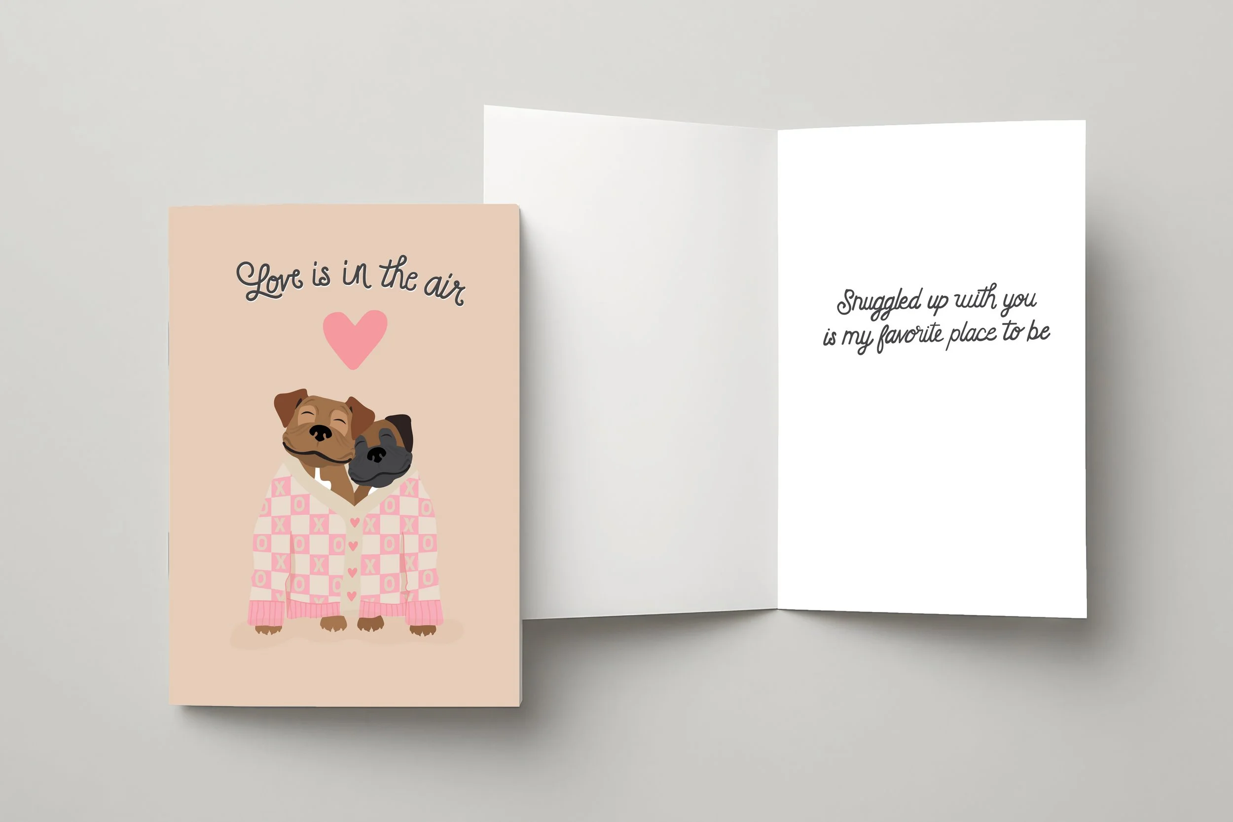 Valentine's Day greeting card featuring two cuddling dogs under pink checkered blanket with text 'Love is in the air' and inside message 'Snuggled up with you is my favorite place to be
