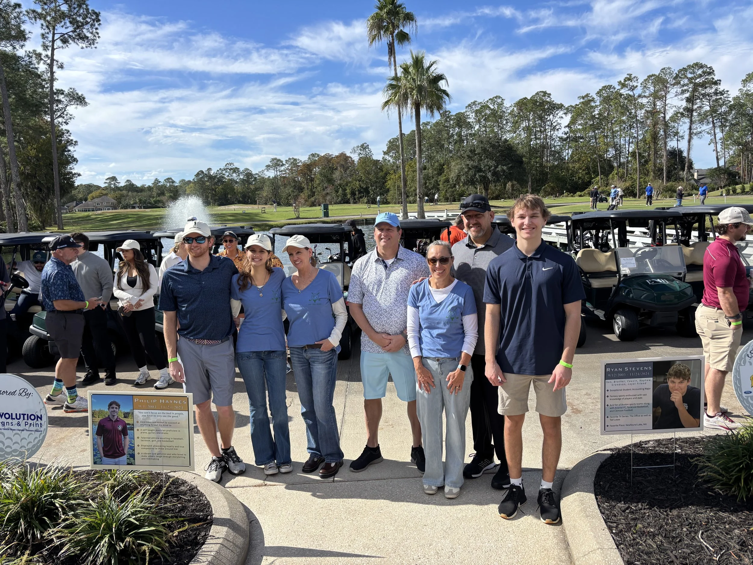 Philip and Ryan's family standing in front of golf carts on a golf course, with trees and a fountain in the background, on a sunny day, for the golf tournamnet.