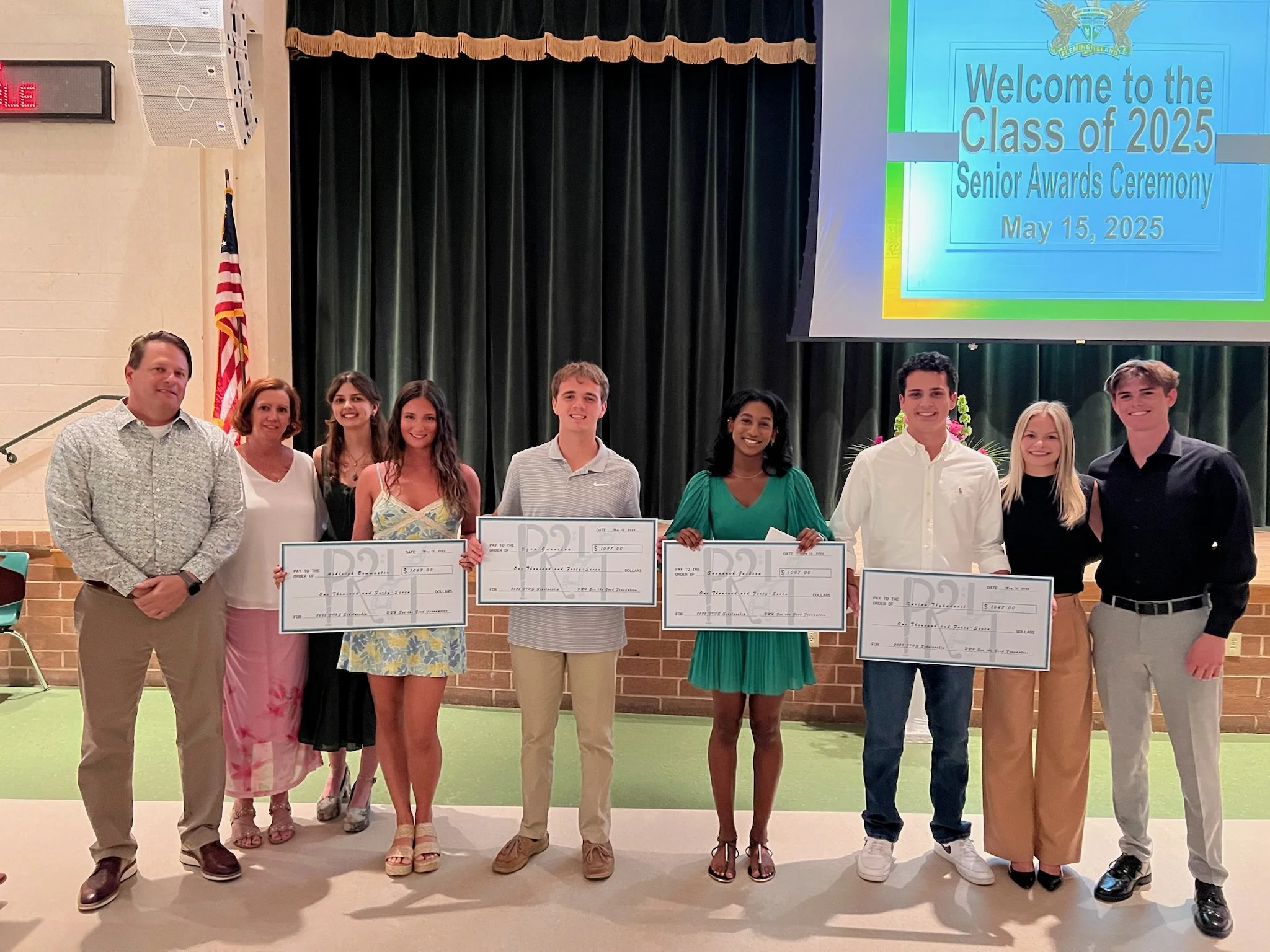 A group of nine people standing on a stage during a high school senior awards ceremony. Four students are holding oversized checks, and a presentation screen in the background displays a message welcoming the class of 2025.