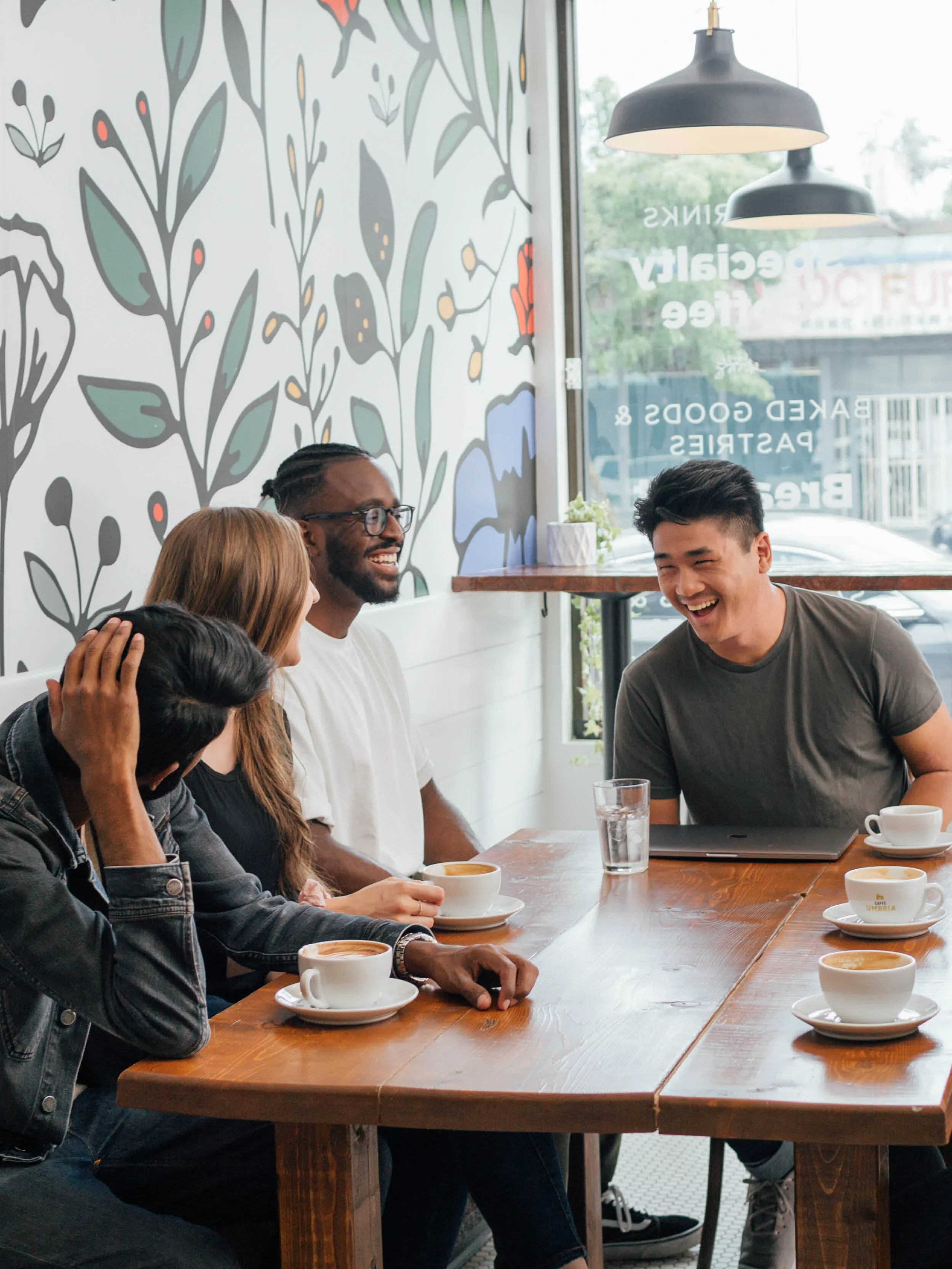 A group of people smiling and chatting at a round table in a cozy cafe or restaurant. The table has cups of coffee, glasses of water, a teapot, and notebooks.