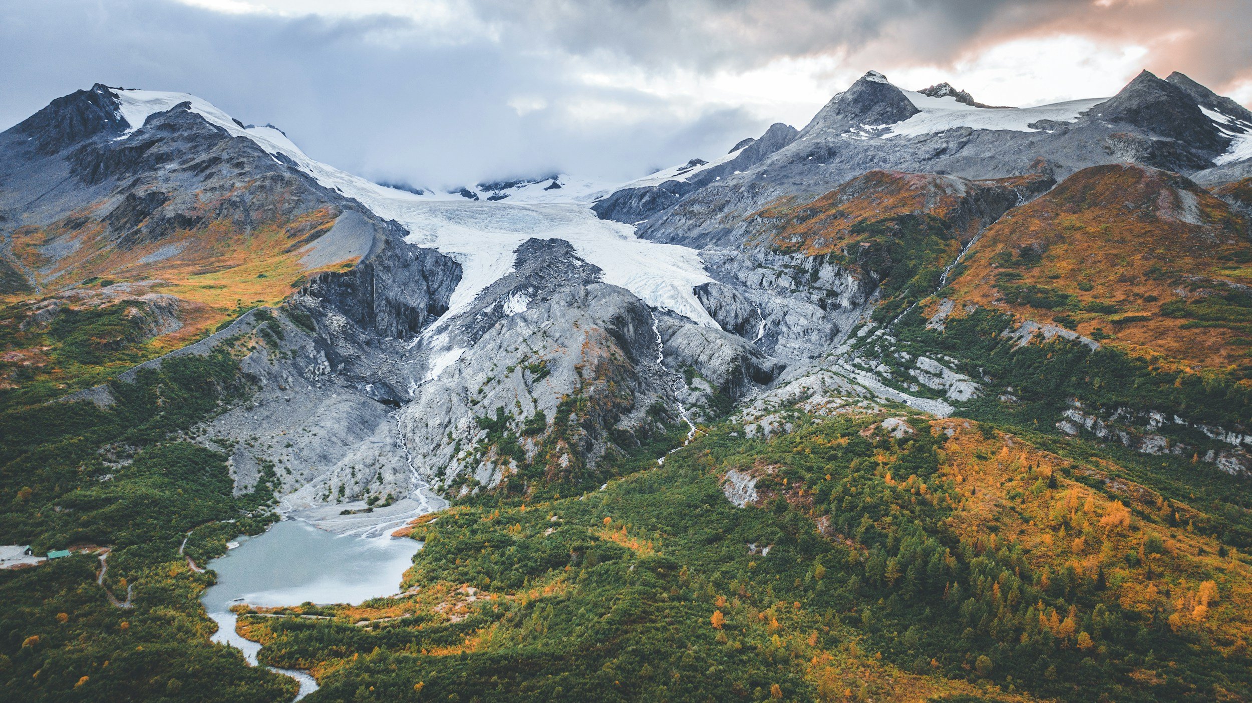 Mountain landscape with a glacier, surrounded by rocky peaks and autumn-colored vegetation. A small lake is visible in the foreground.