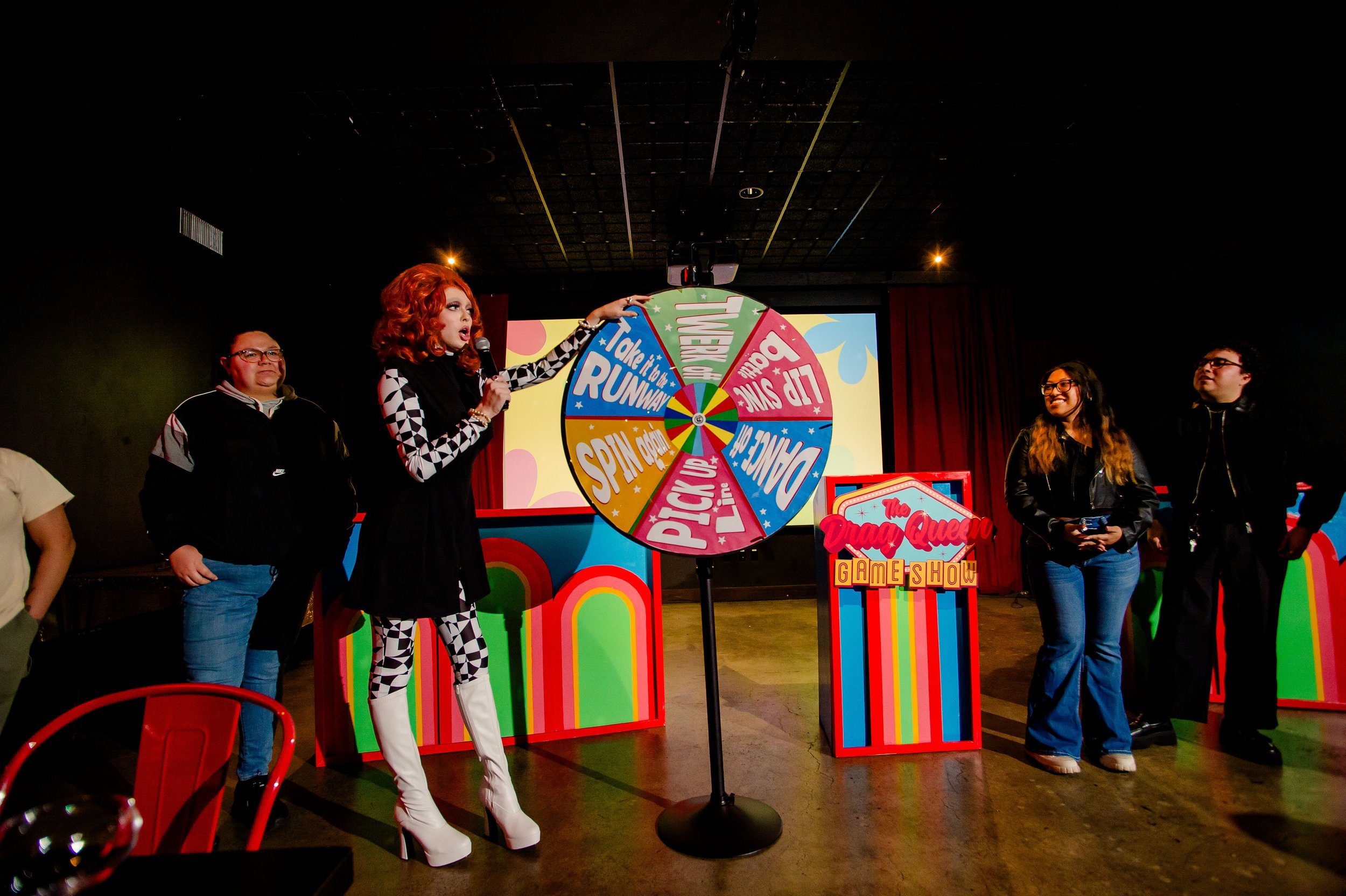 Group of people at a drag queen game show with a colorful prize wheel on stage.