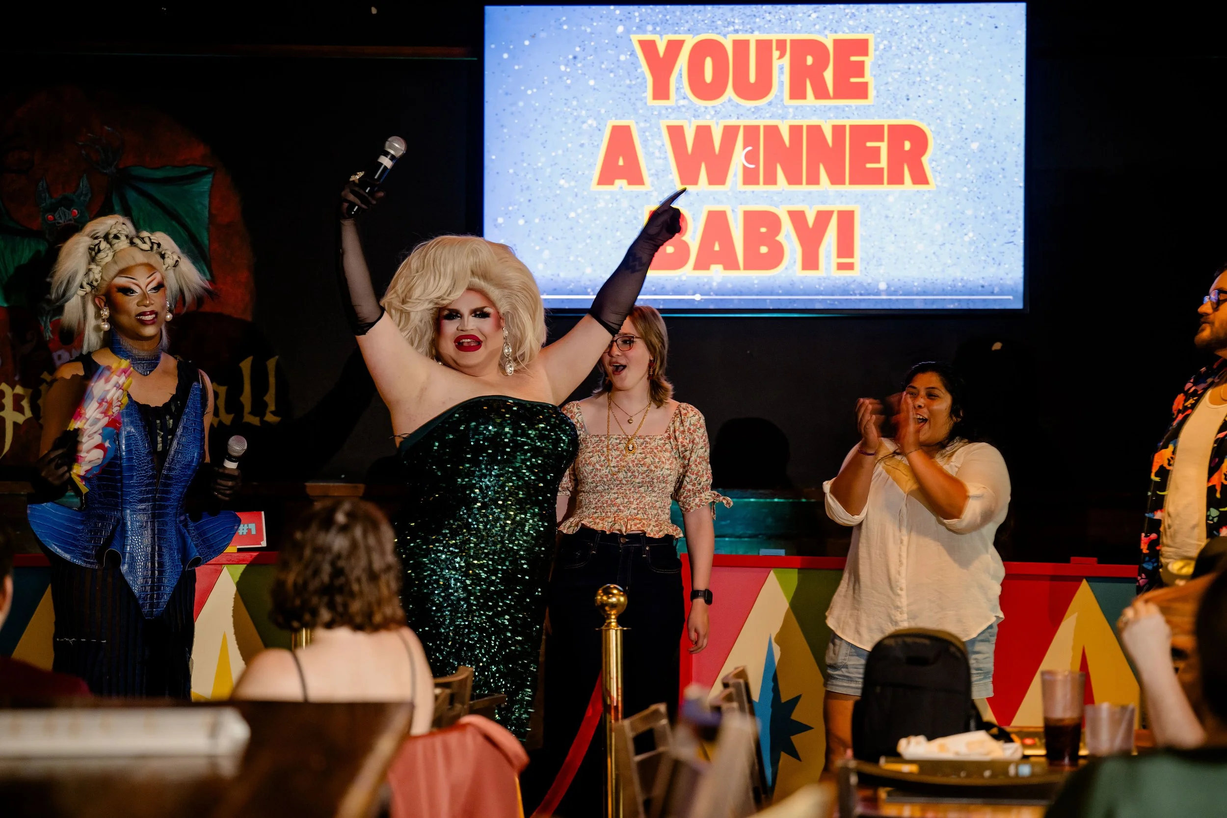 Drag queens on stage celebrating a victory at a bingo event with a sign that reads 'You're a Winner Baby!' in the background at the Drag Queen Game Show