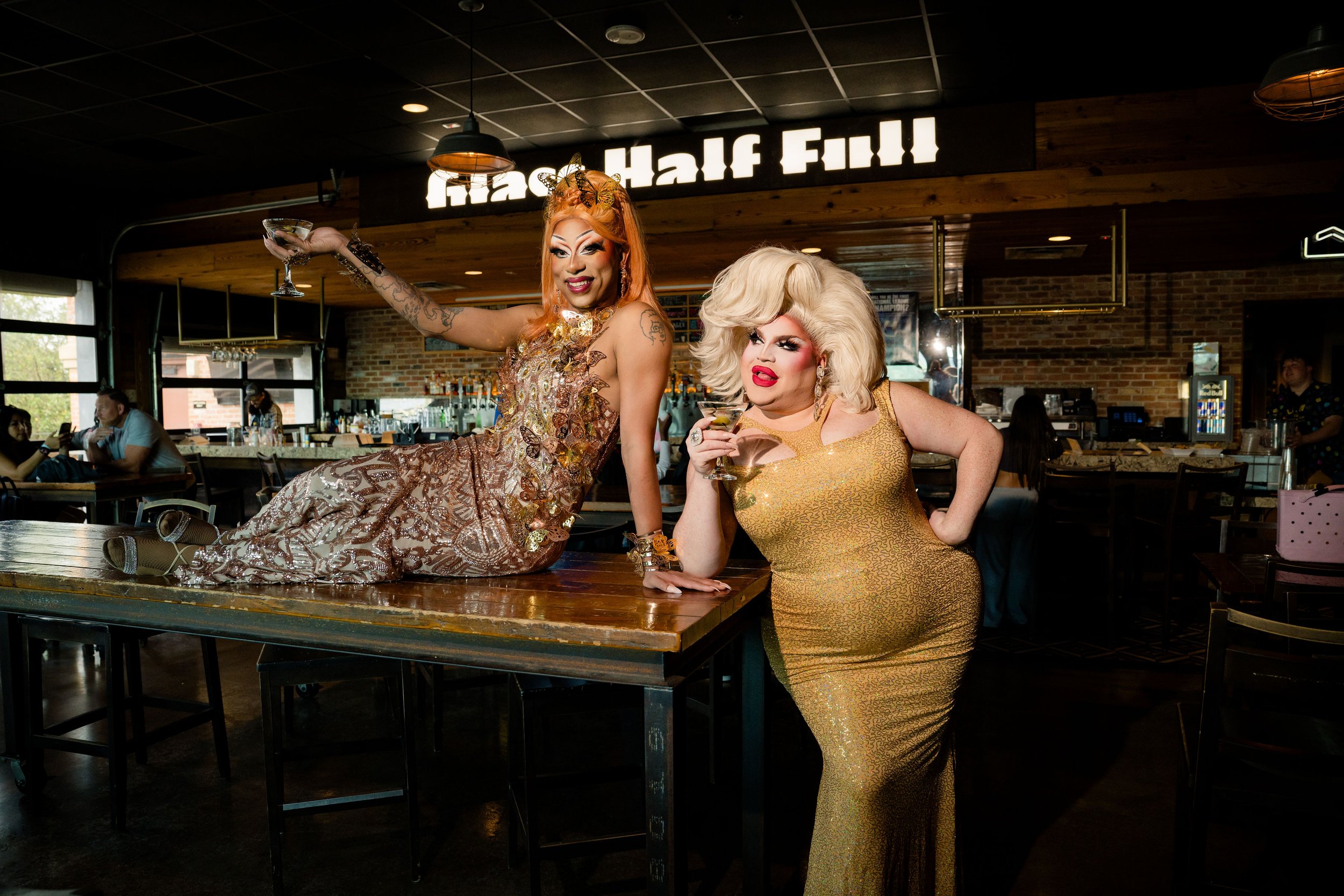Two drag queens posing and smiling at a bar, one sitting on the bar with an arm raised holding a martini glass, the other standing next to her with a glass. The bar has a sign that reads 'Half Fill' in the background, the Cedars bar.