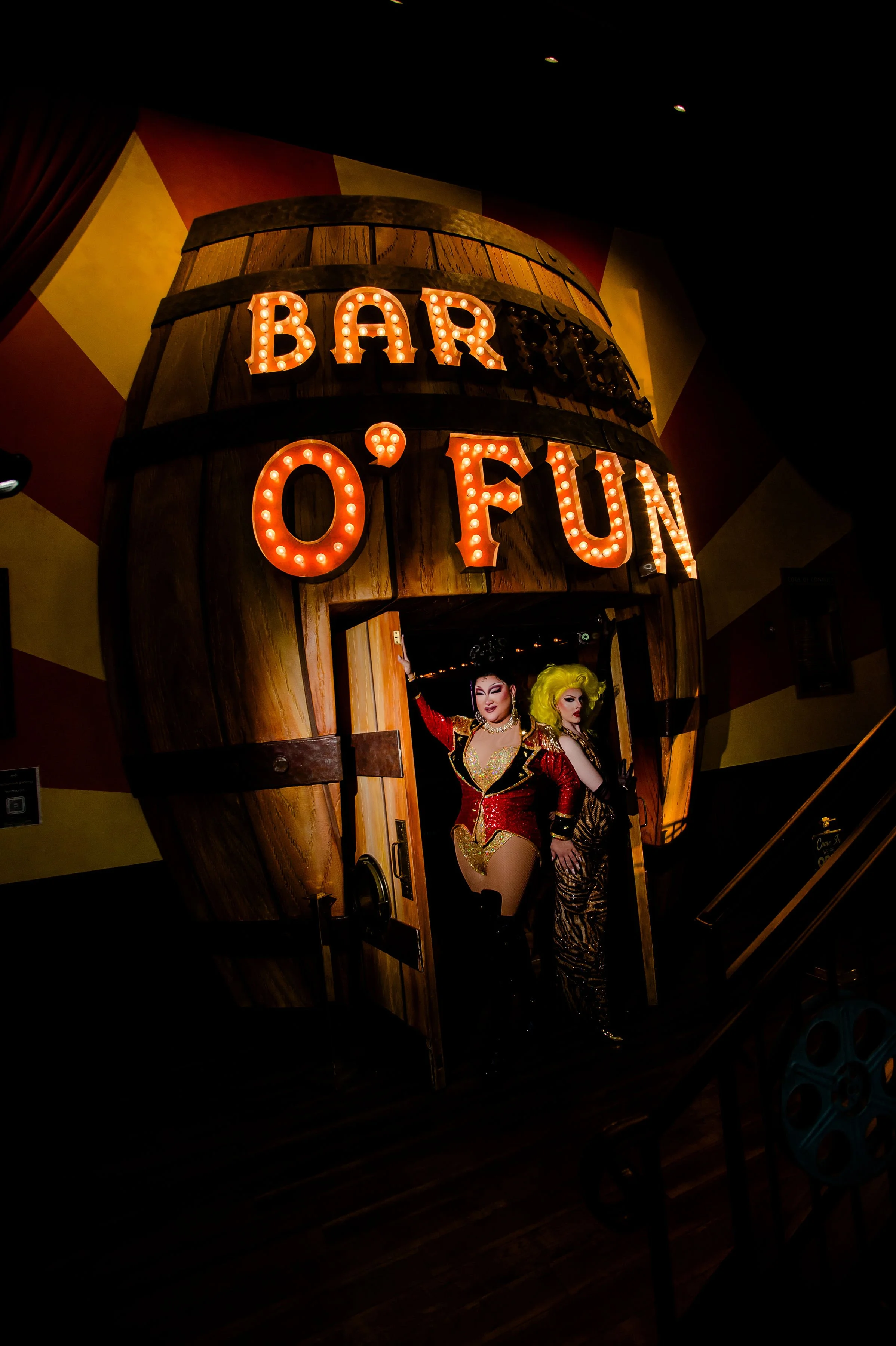 Two drag performers on stage in a circus-themed setting, one standing and holding a whip, the other kneeling with vibrant yellow hair, surrounded by colorful retro decor and signs.