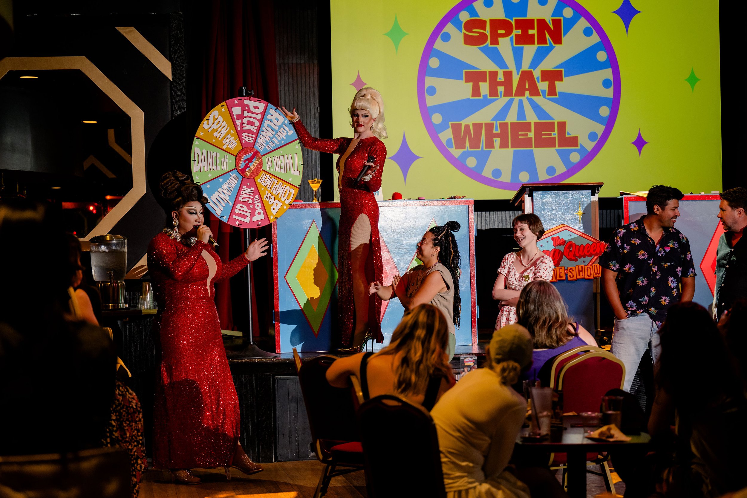 People participating in a live game show with a brightly colored wheel and stage, hosted by two performers in red costumes.