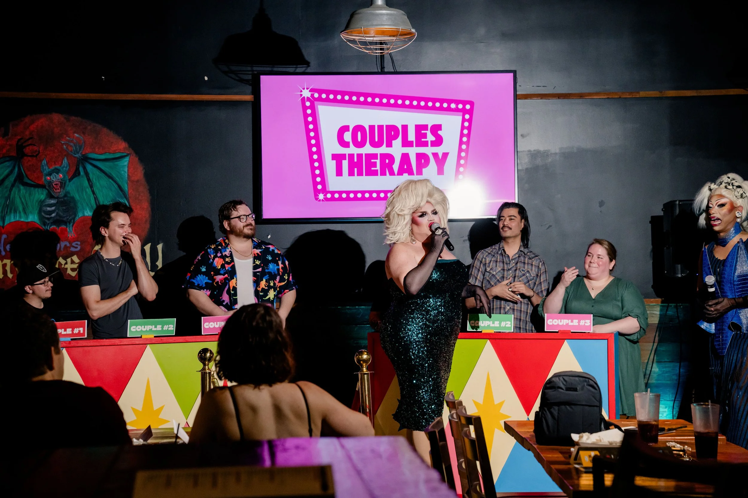 A drag queen in a sparkling black dress performing on stage at the Drag Queen Game Show, with a large pink sign behind her reading 'COUPLES THERAPY' and several contestants sitting at a table.