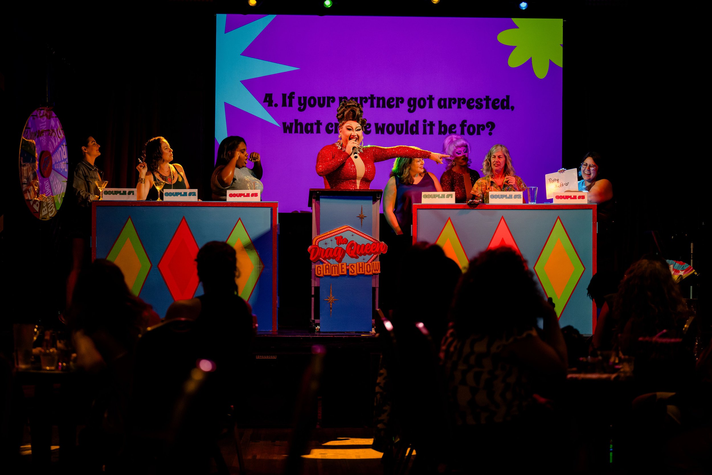 People participating in a game show hosted by a drag queen, with colorful podiums and a question projected on a screen.