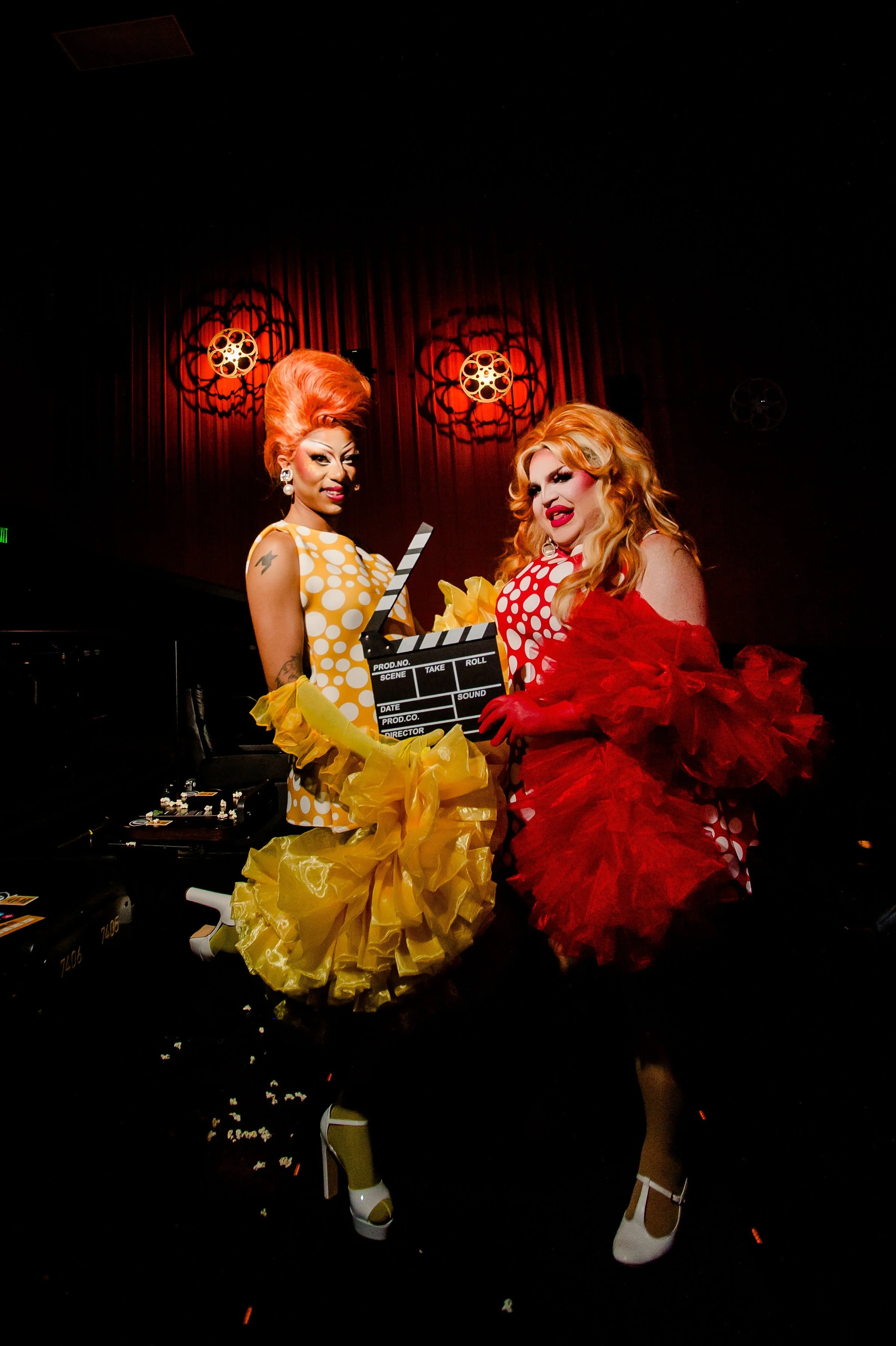 Two drag queens performing on stage, dressed in vintage-style polka dot dresses with ruffled skirts, wearing high heels and glam makeup, holding a movie clapperboard, with dramatic lighting and red curtains in the background.