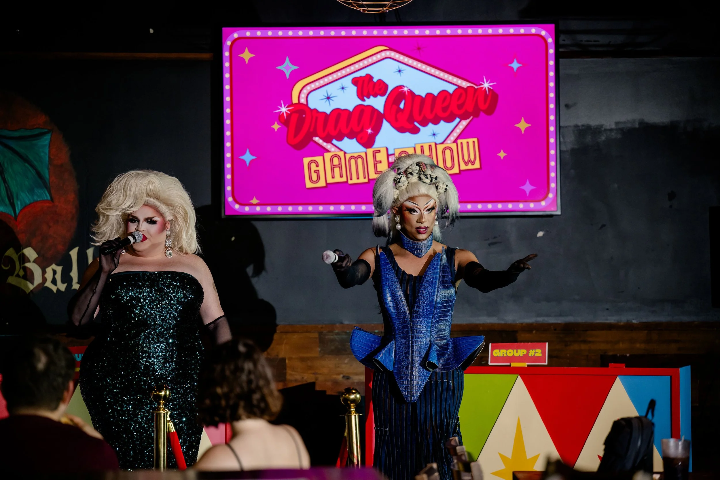 Two drag queens performing on stage at the Drag Queen Game Show with a bright pink sign behind them reading 'The Drag Queen Game Show'. The stage is decorated with colorful geometric shapes and a colorful audience watches the performance.
