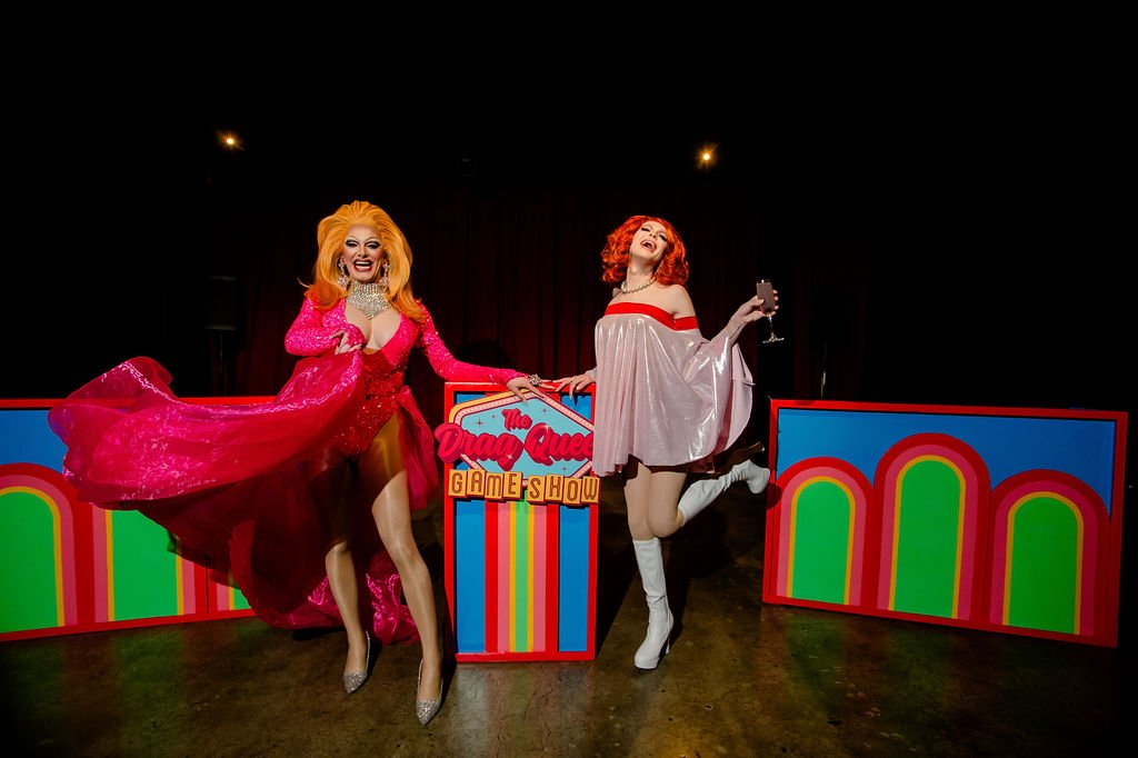 Two drag queens in colorful outfits on stage with a "Drag Queen Game Show" sign.