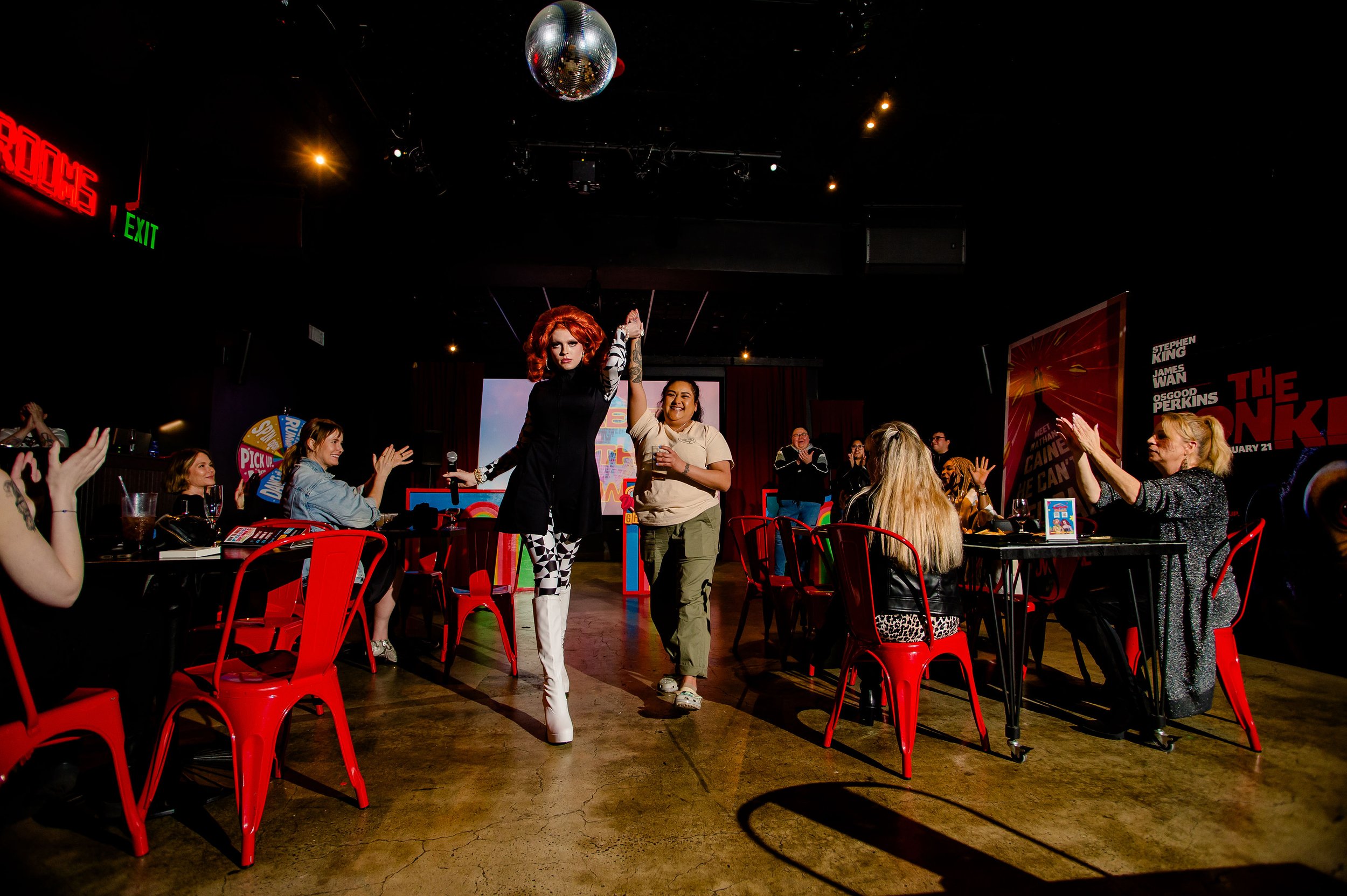 Performers on stage during a show with audience clapping, red chairs, and a disco ball overhead.