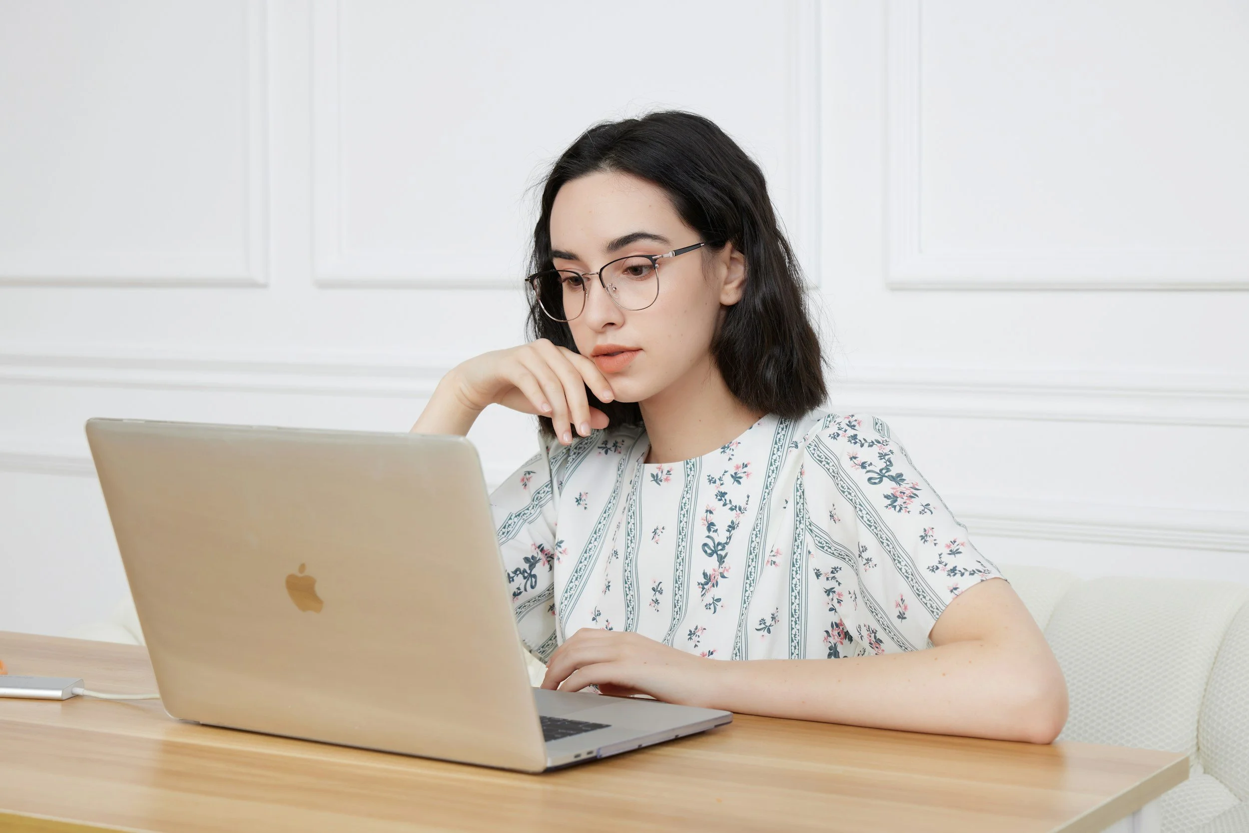 A young woman with black hair, glasses, and light skin sitting at a wooden table, looking at an open silver MacBook computer.