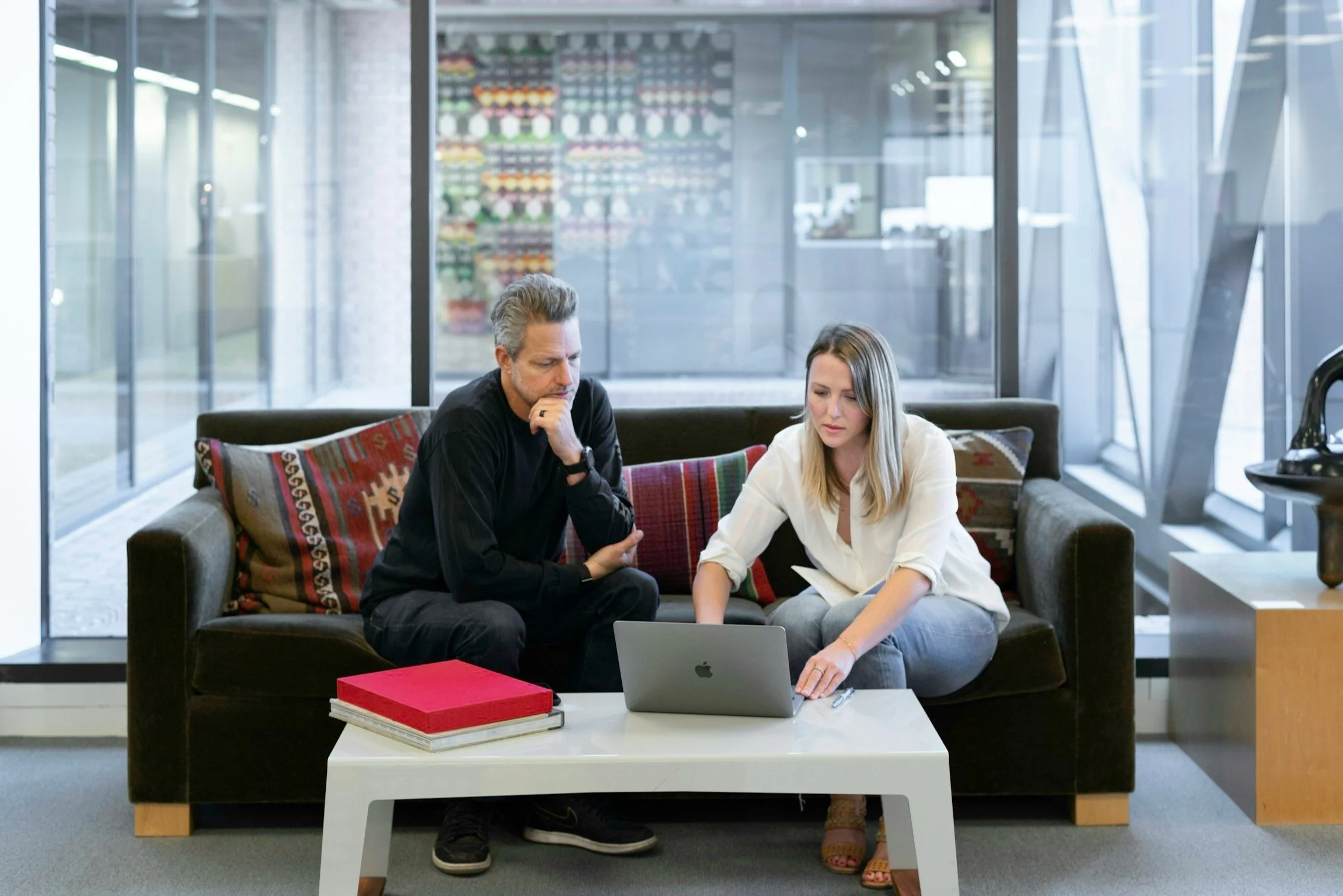 Two people, a man and a woman, sitting on a black sofa in a modern office, looking at a laptop on a white coffee table in front of them. The man has gray hair and is wearing a black long-sleeve shirt, while the woman has blonde hair and is wearing a white blouse. They appear to be discussing something on the laptop.