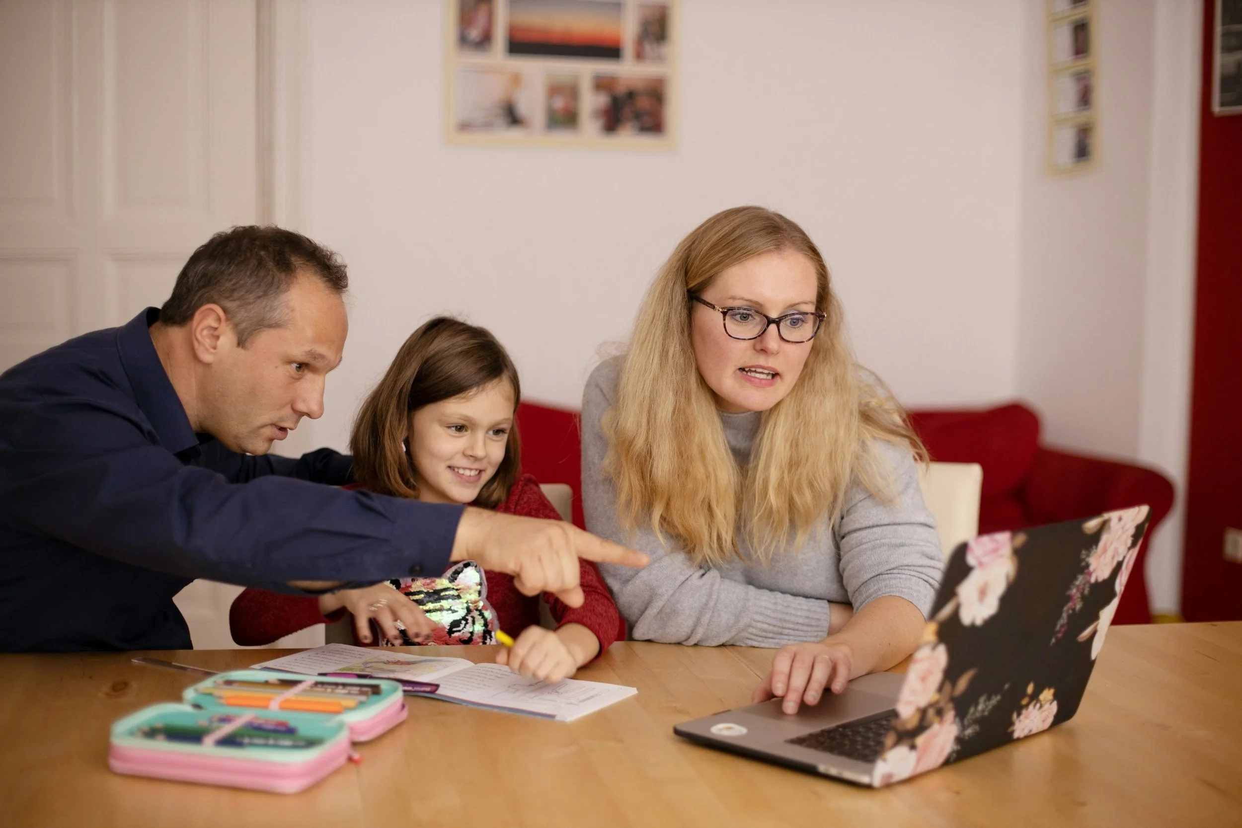 A family with a young girl sits at a table. The father is pointing at a laptop screen, the girl is smiling, and the mother is looking at the screen. On the table are a pencil case and an open notebook.