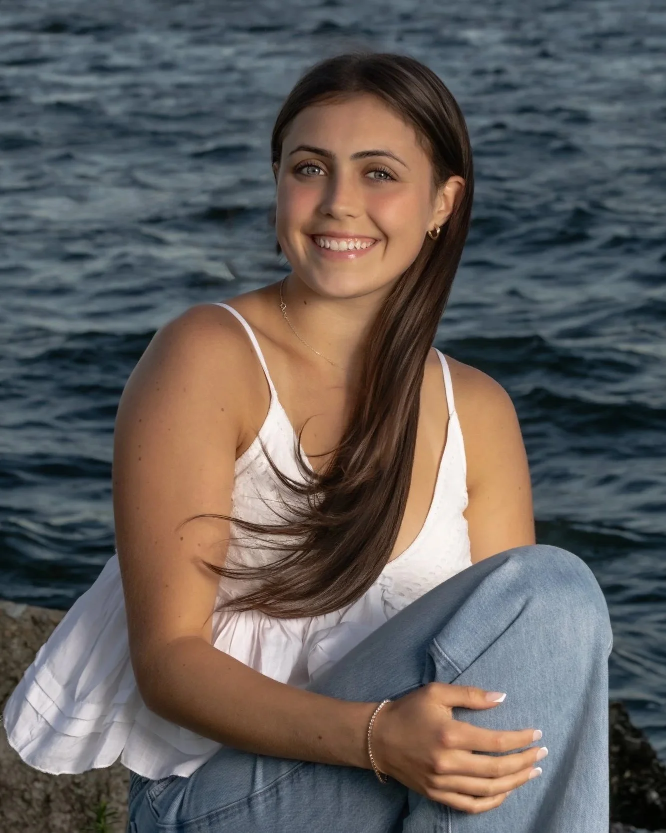 High school senior girl in a white top and jeans posing on the rocky shore at Rocky Point State Park in Warwick, Rhode Island.
