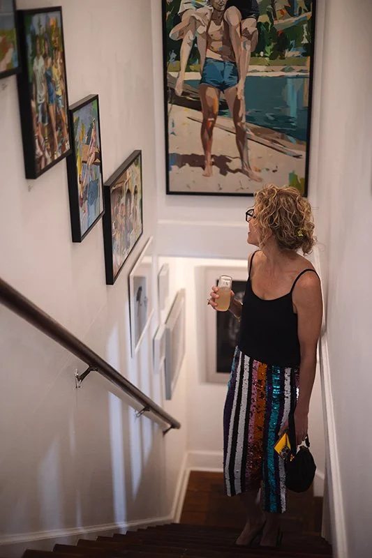 Catherine Blount, founder of Candler Creative, stands on a staircase landing in an art gallery, holding a drink in one hand and a small purse in the other, looking at paintings on the wall.