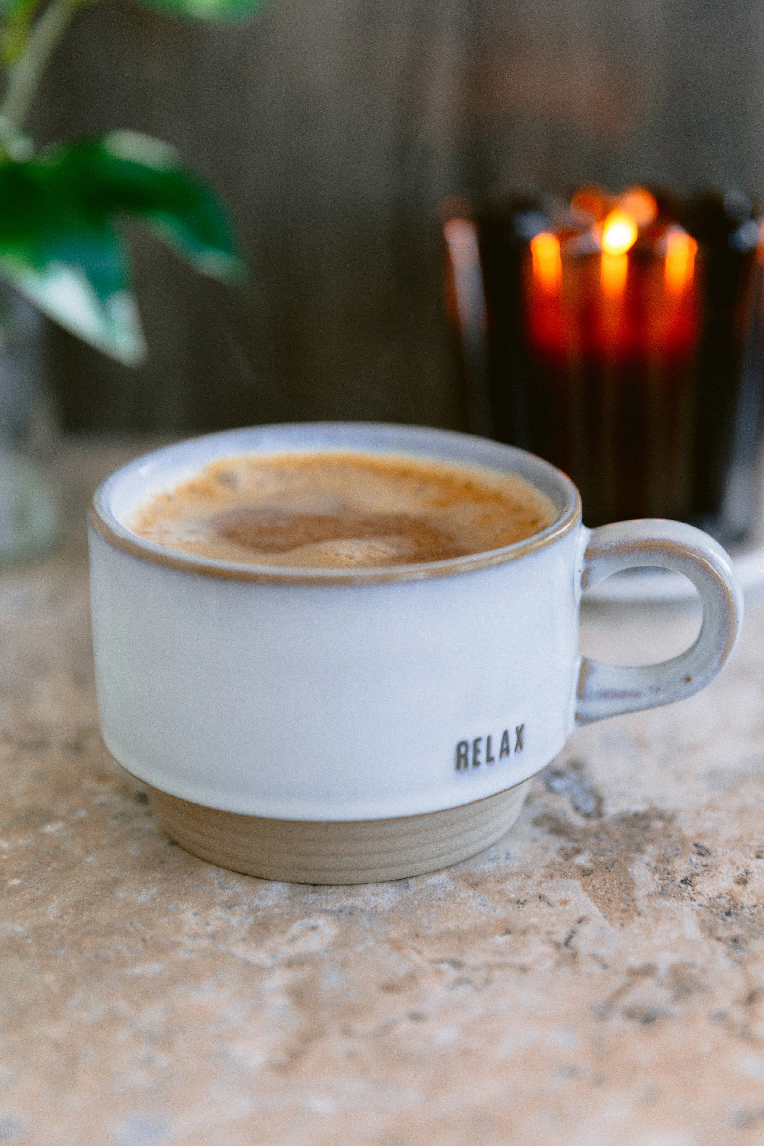 A white ceramic mug with the word 'RELAX' written on it, filled with coffee or a similar beverage, placed on a textured surface with a candle in the background.