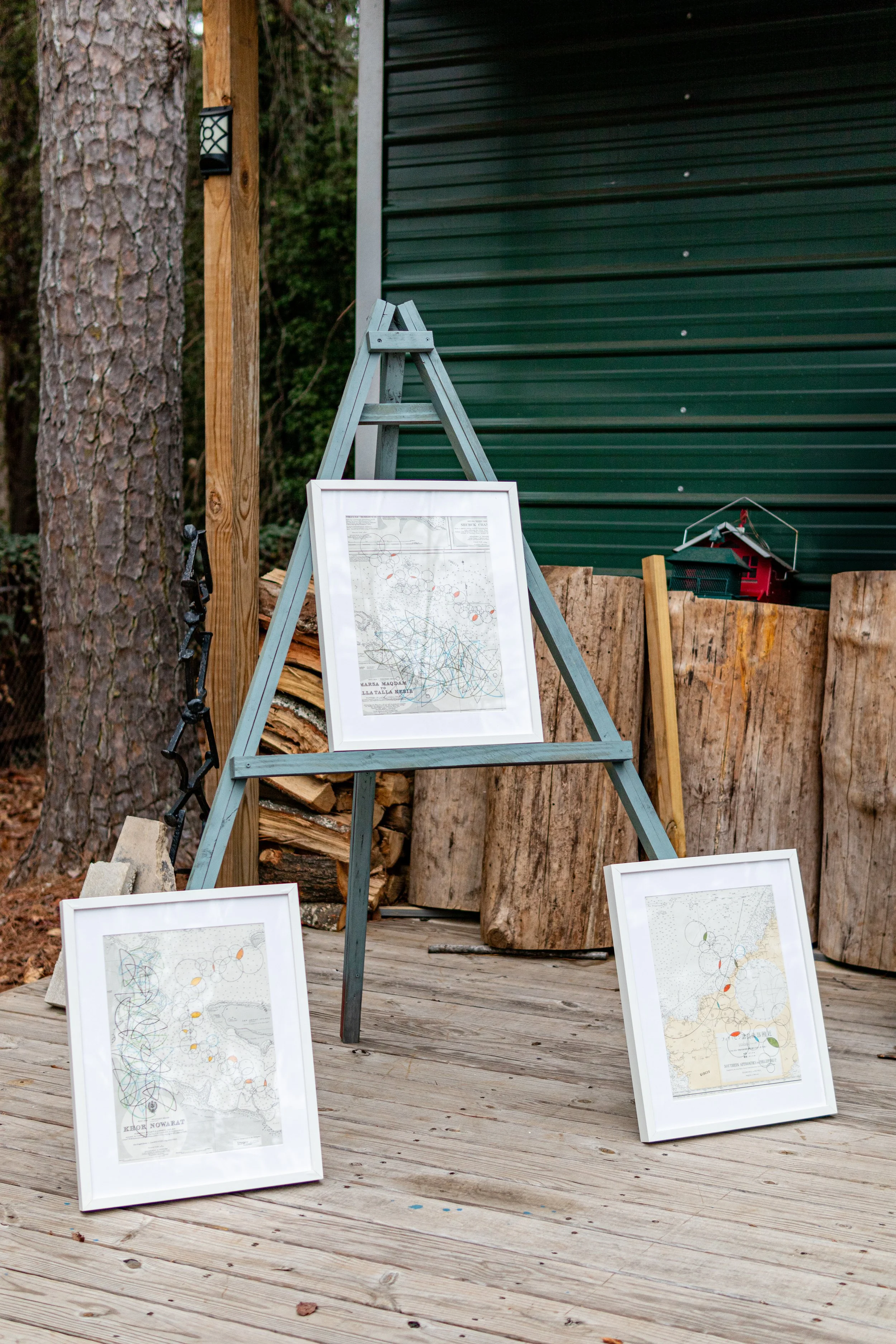 Three framed abstract map artworks on a wooden deck, with logs and a green metal building in the background.
