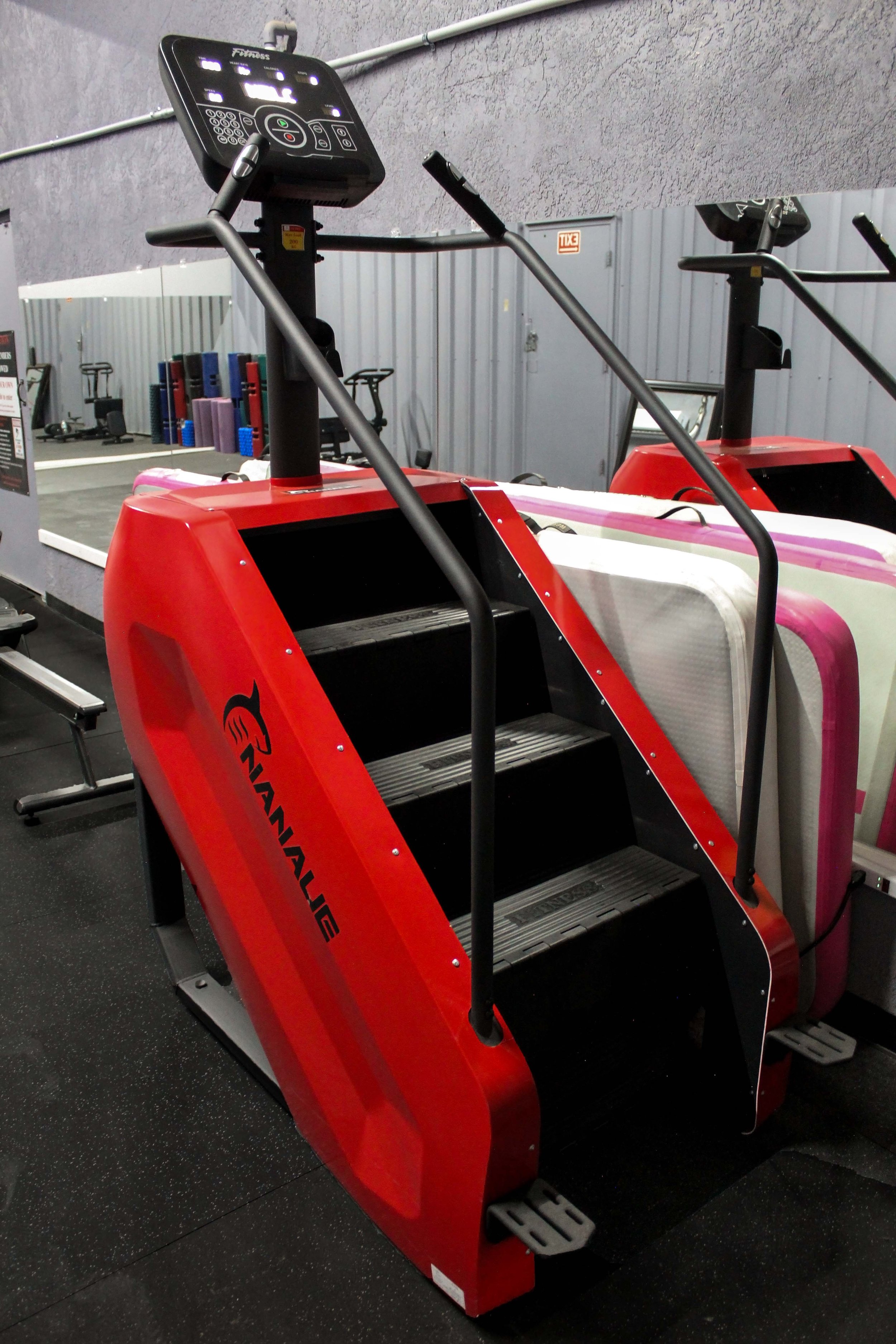 Red Nanaji stair climbing machine with a digital console, positioned on rubber flooring in a gym with mirrored walls and nearby equipment.