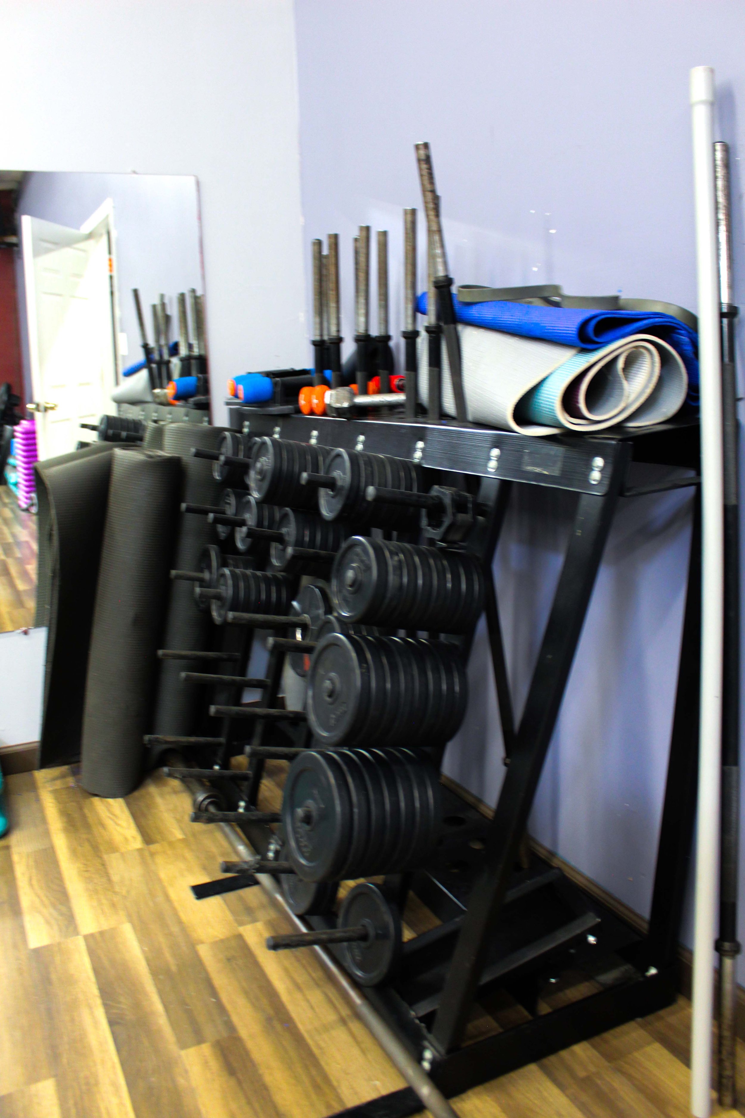 Rack of custom weight barbells with adjustable plates, alongside yoga mats and workout accessories, set against a light purple wall on a wooden floor.