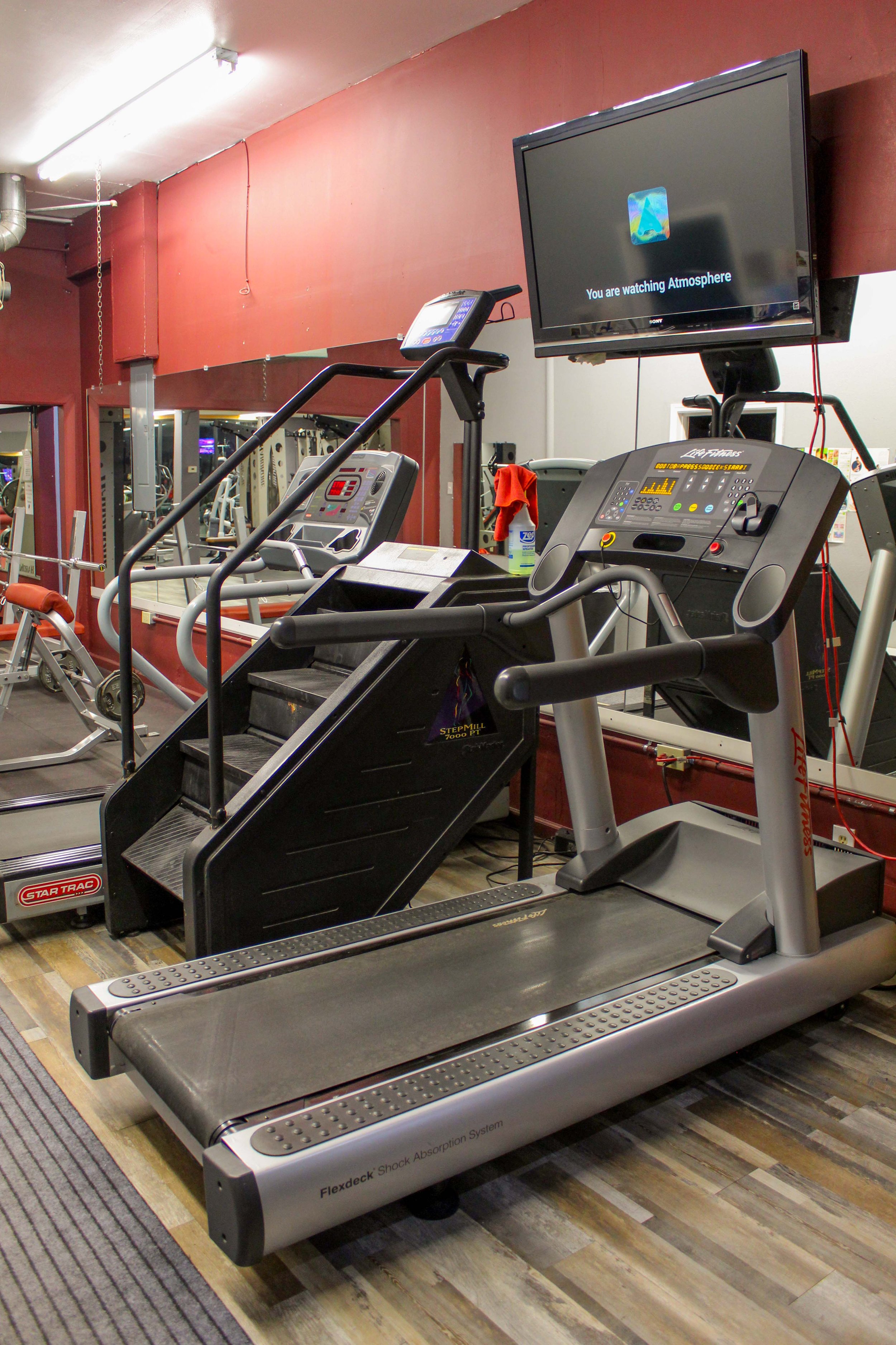 StairMaster StepMill and Life Fitness treadmill positioned on a wooden-style floor, with a wall-mounted TV displaying 'You are watching Atmosphere' in a gym setting.