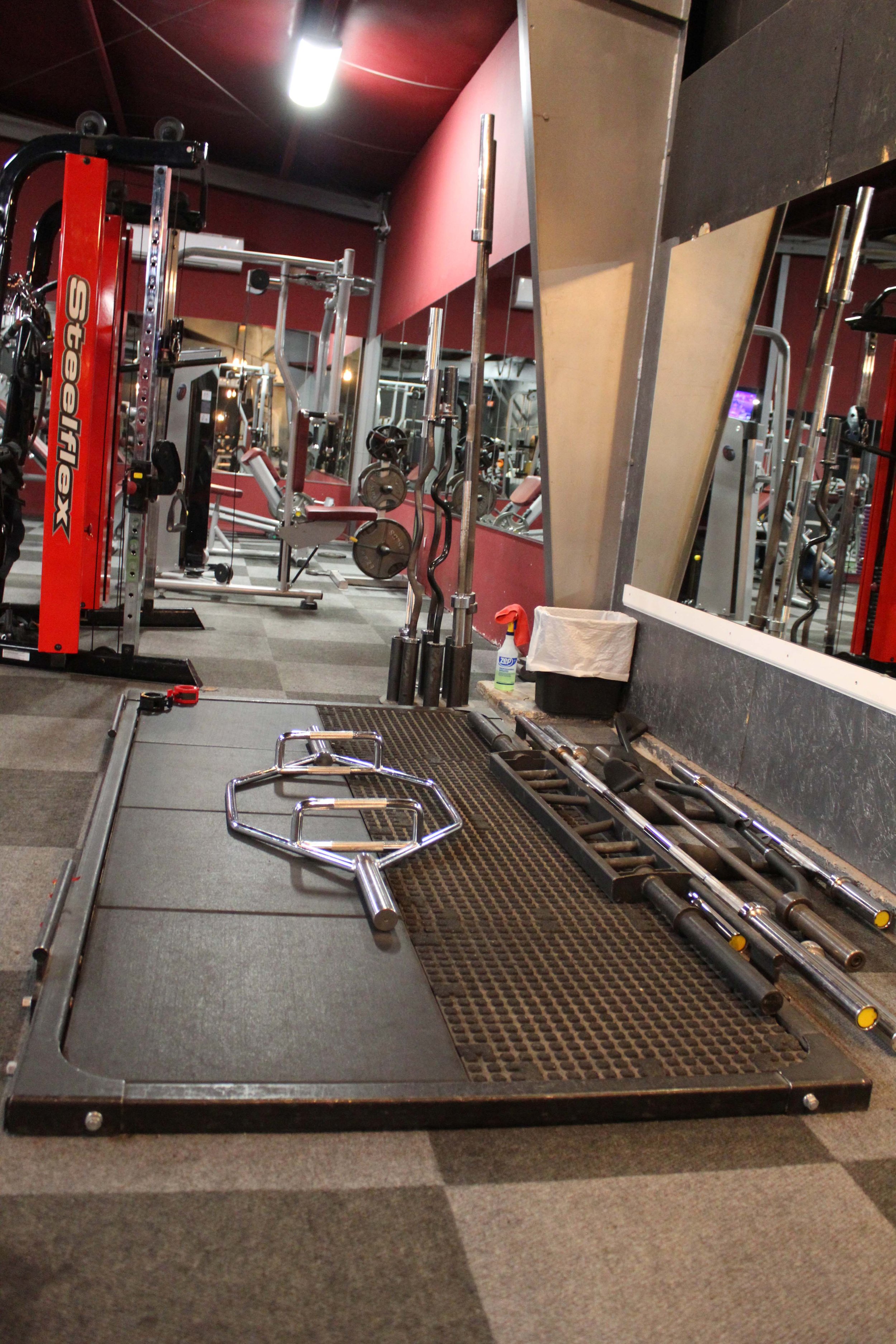 Deadlift platform with hex bar, barbell bars, and weights neatly arranged, located in a gym with mirrored walls and red SteelFlex equipment in the background.