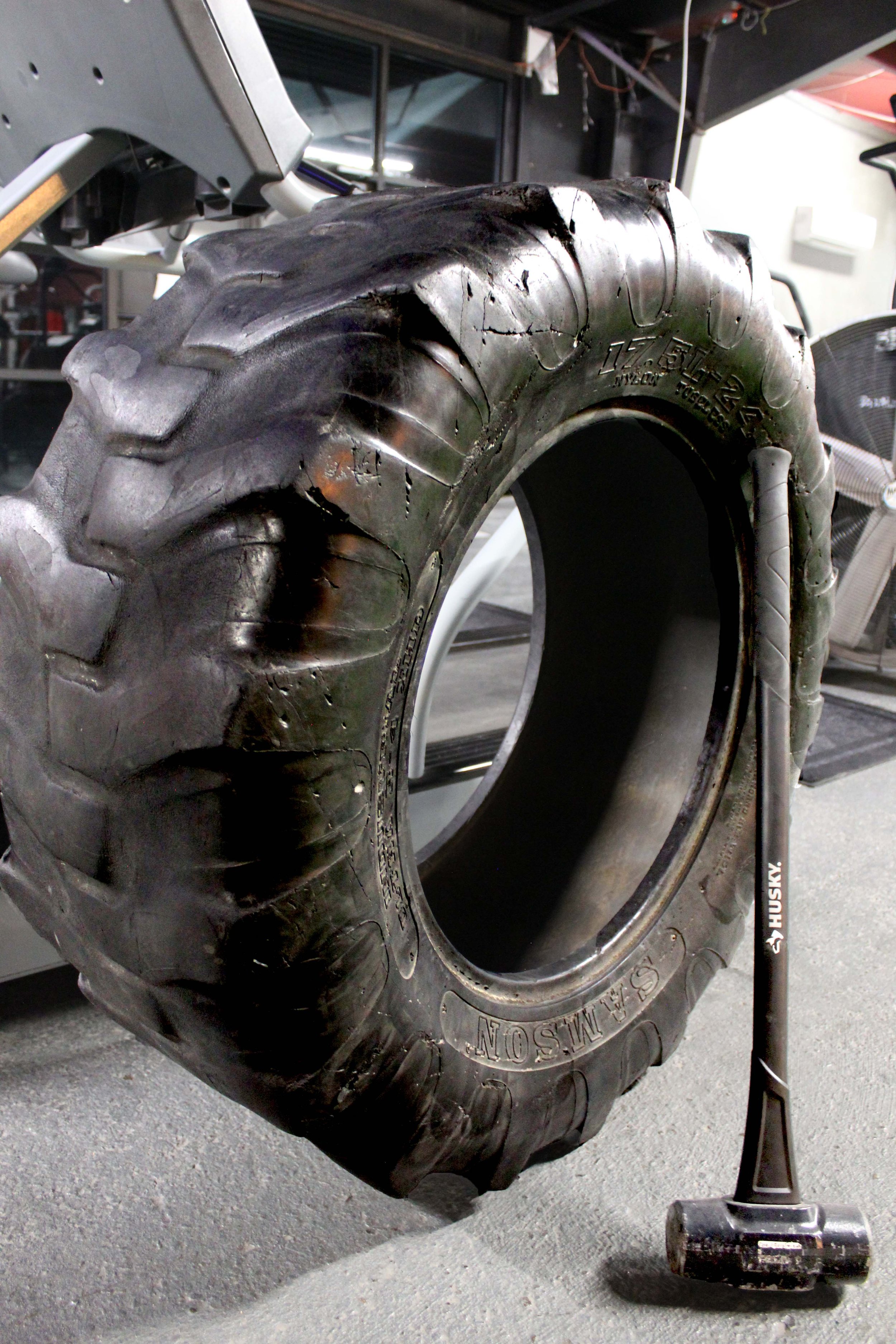 Large tractor tire used for fitness training, paired with a Husky sledgehammer, positioned on a rubber gym floor.
