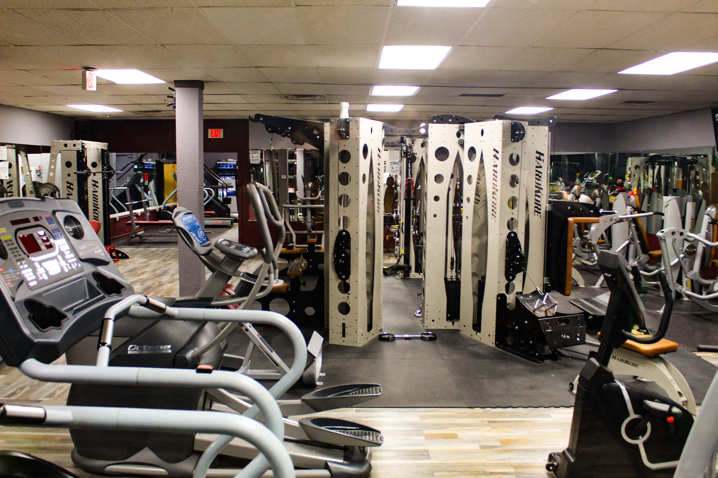 Wide-angle view of a gym interior showcasing various fitness equipment, including elliptical machines in the foreground and a multifunctional jungle gym station at the center, all surrounded by mirrors and a clean, well-lit environment.