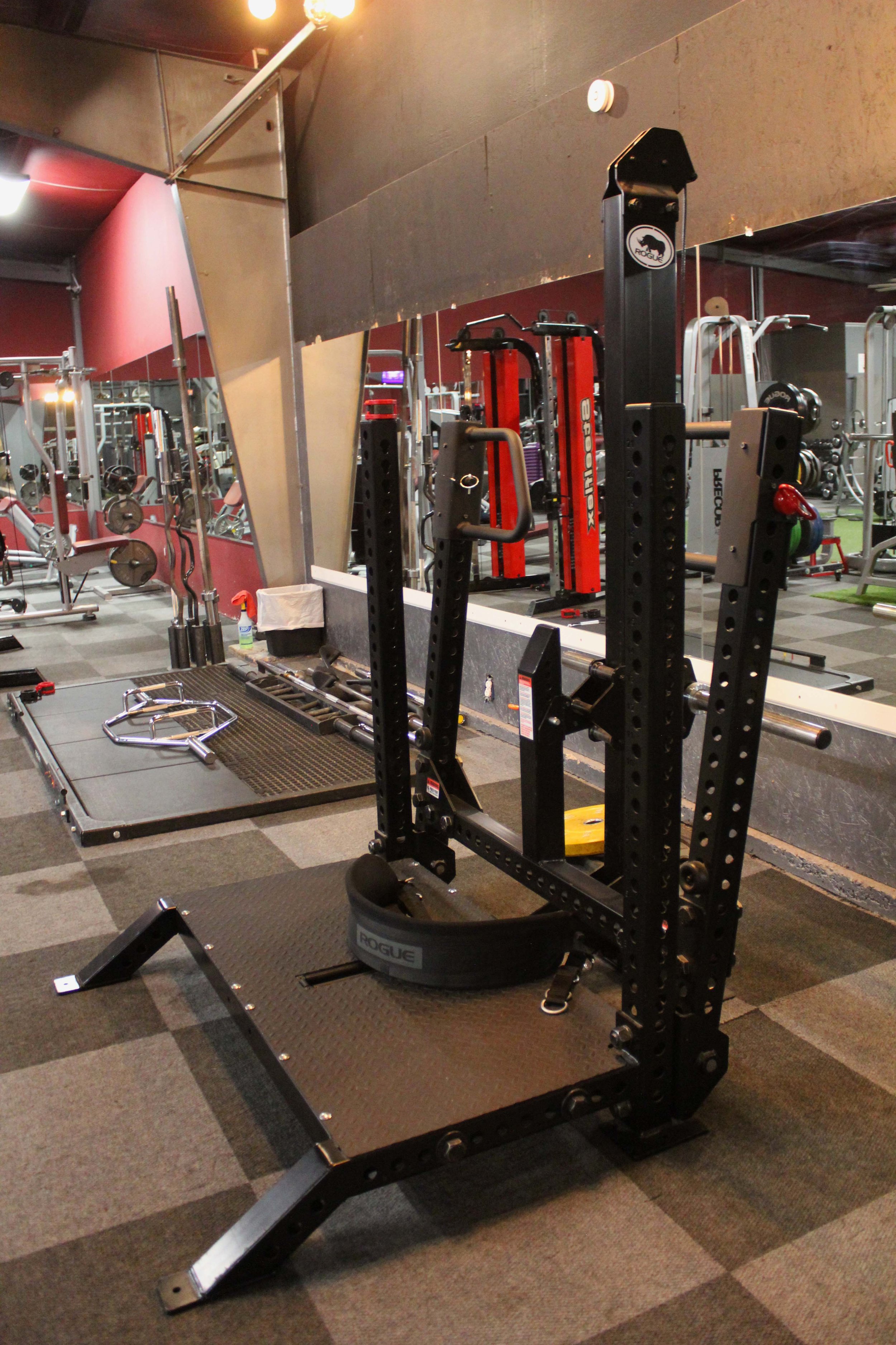 Rogue specialty squat machine with a sturdy black frame and padded belt, set in a gym with mirrored walls and nearby barbell equipment.