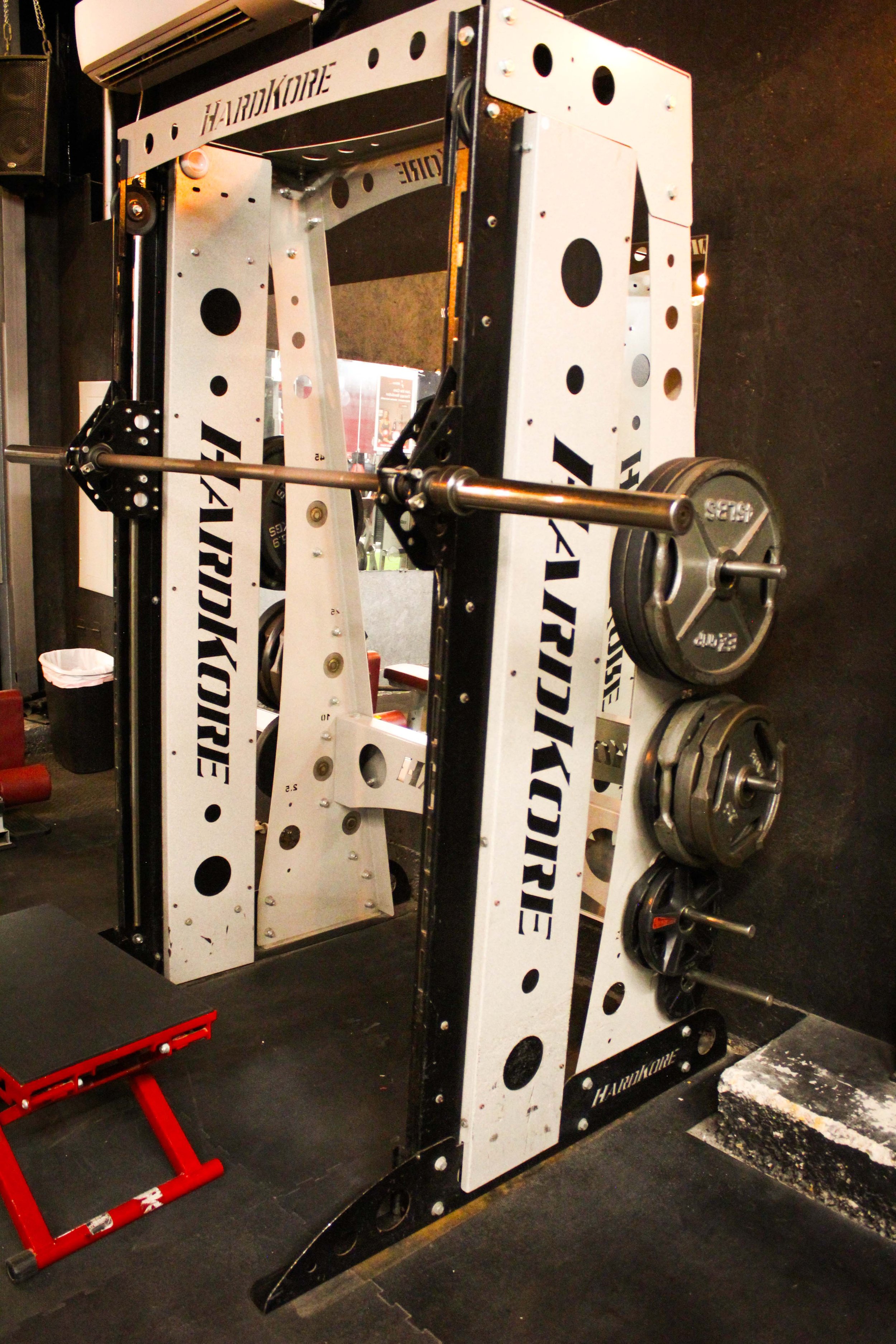 Inclined Smith machine with a barbell loaded with weight plates, featuring a sturdy black-and-white HardKore frame in a gym setting.
