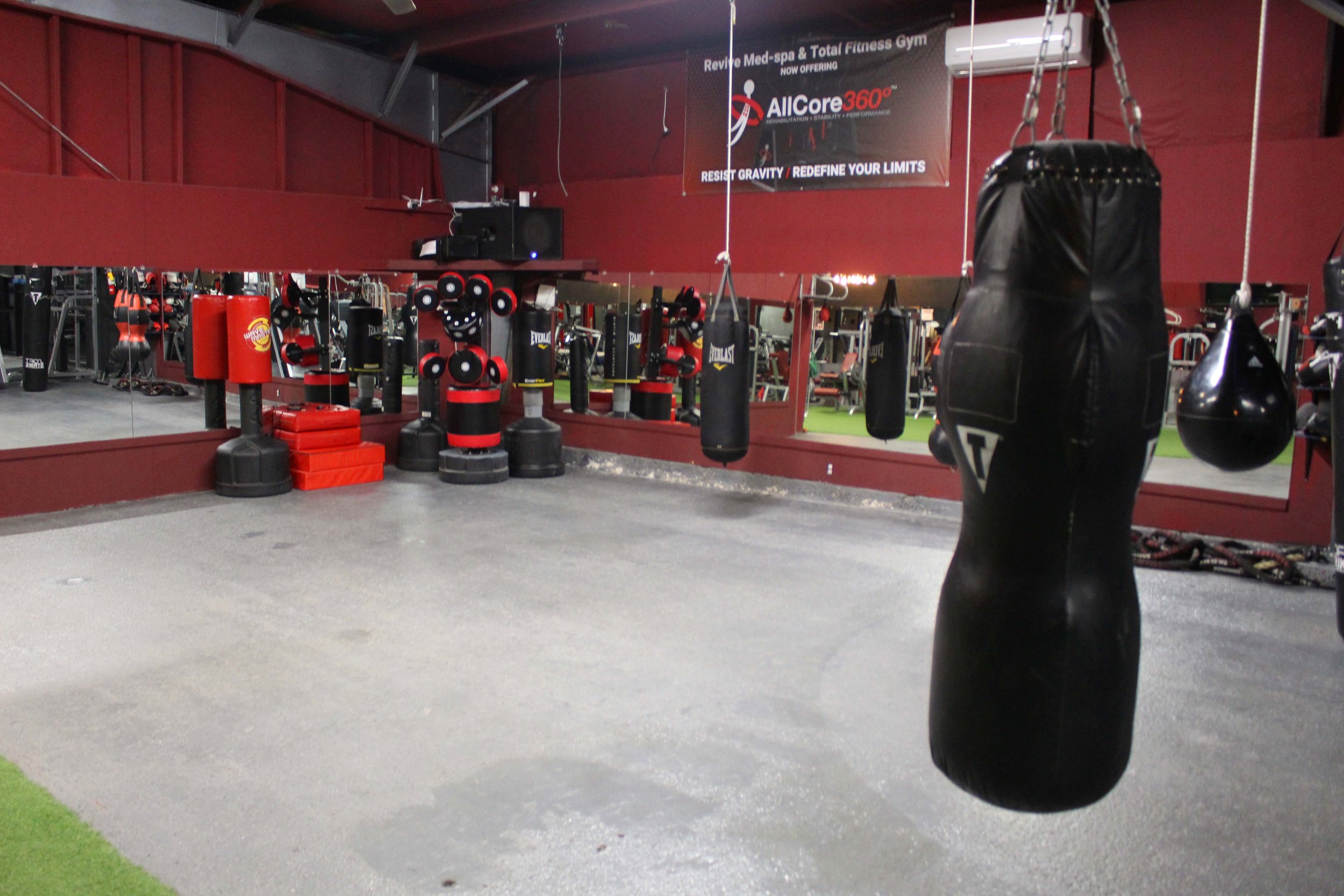 Boxing area in a gym featuring heavy punching bags, red and black training equipment, a mirrored wall