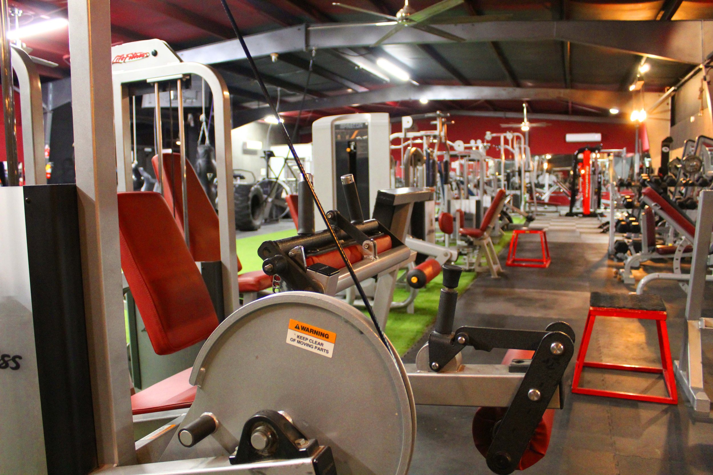 Close-up of a leg workout machine with red seats and visible cables, set in a spacious gym filled with various strength training equipment and machines, under high ceilings with industrial lighting.