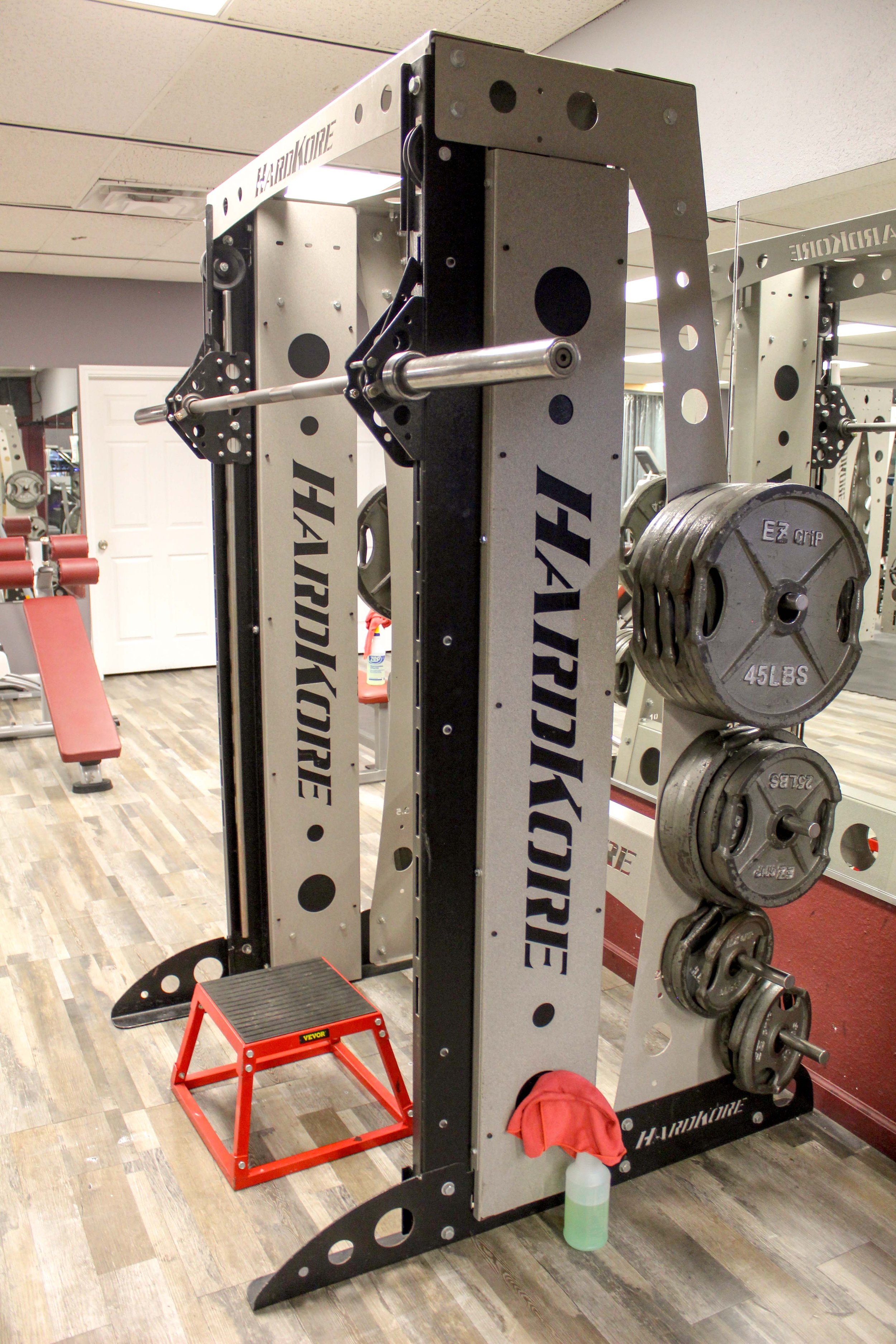 Close-up view of a HardKore straight Smith machine with a loaded barbell and attached weight plates, featuring a red step platform and cleaning supplies at the base. The gym’s clean wood-style flooring and mirrored walls are visible in the background