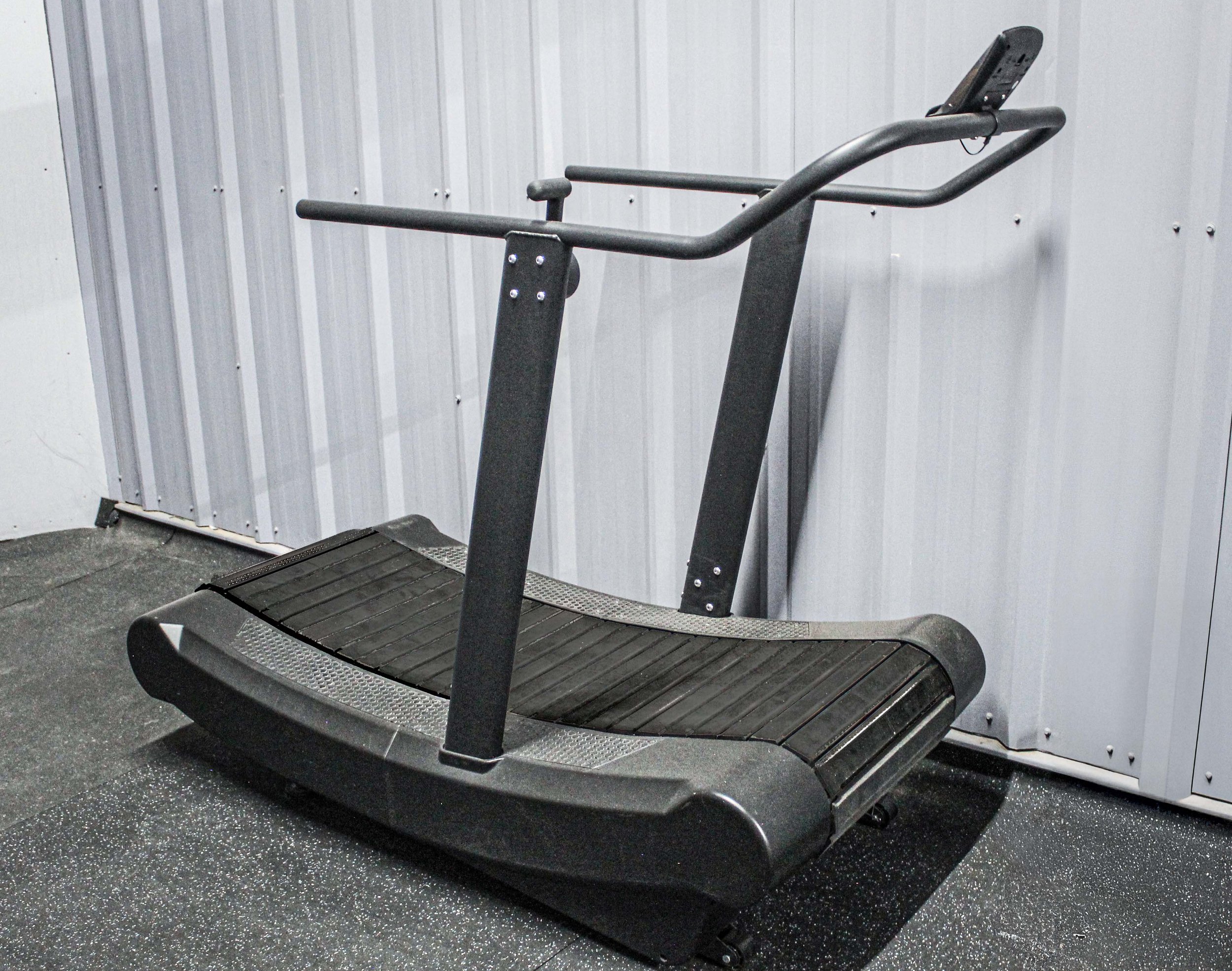 Non-motorized curved treadmill with a sleek black design, positioned against a white metal wall in a gym setting.