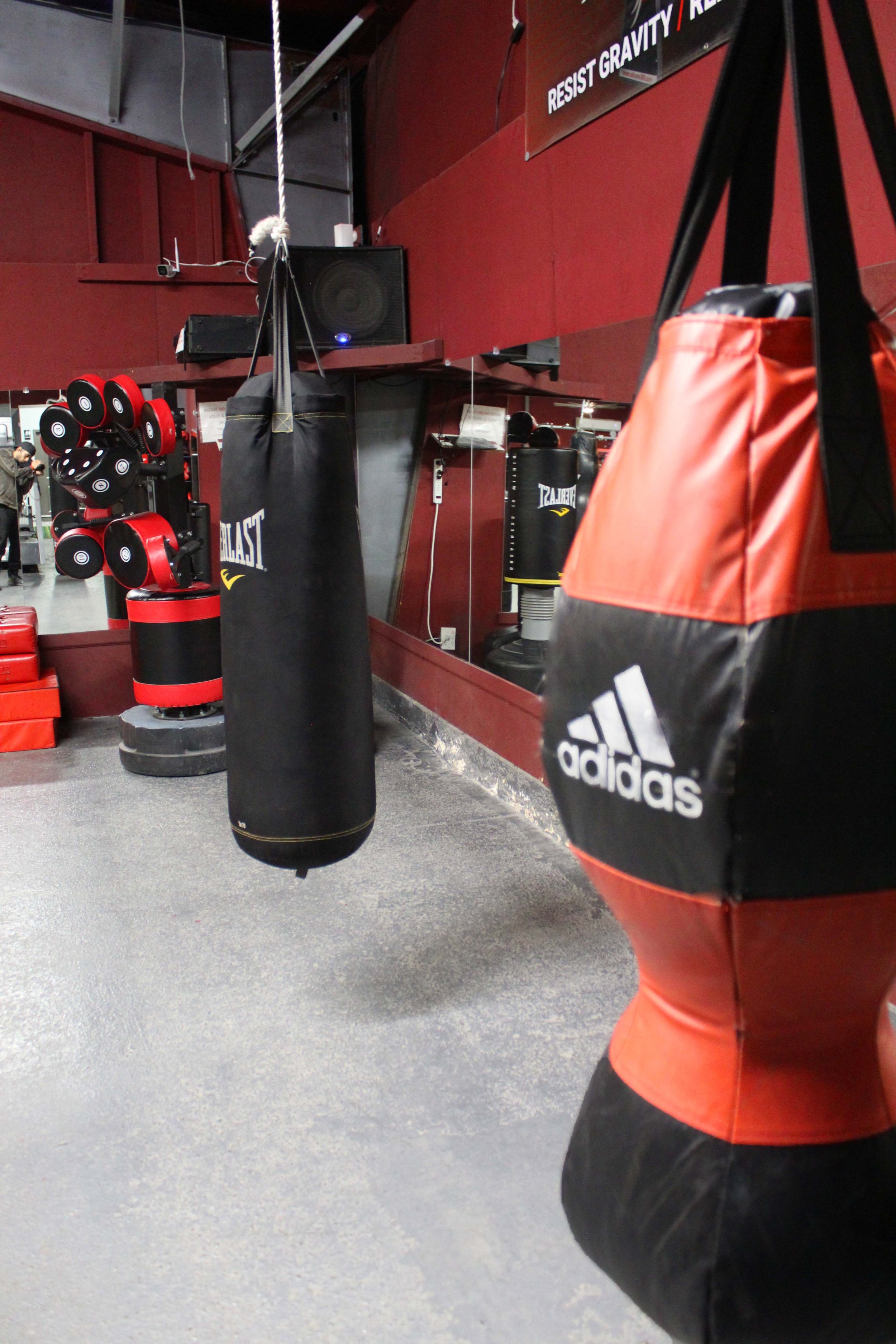 Close-up of Adidas red and black punching bag with an Everlast heavy bag in the background, set in a gym's boxing area featuring red walls and training equipment.