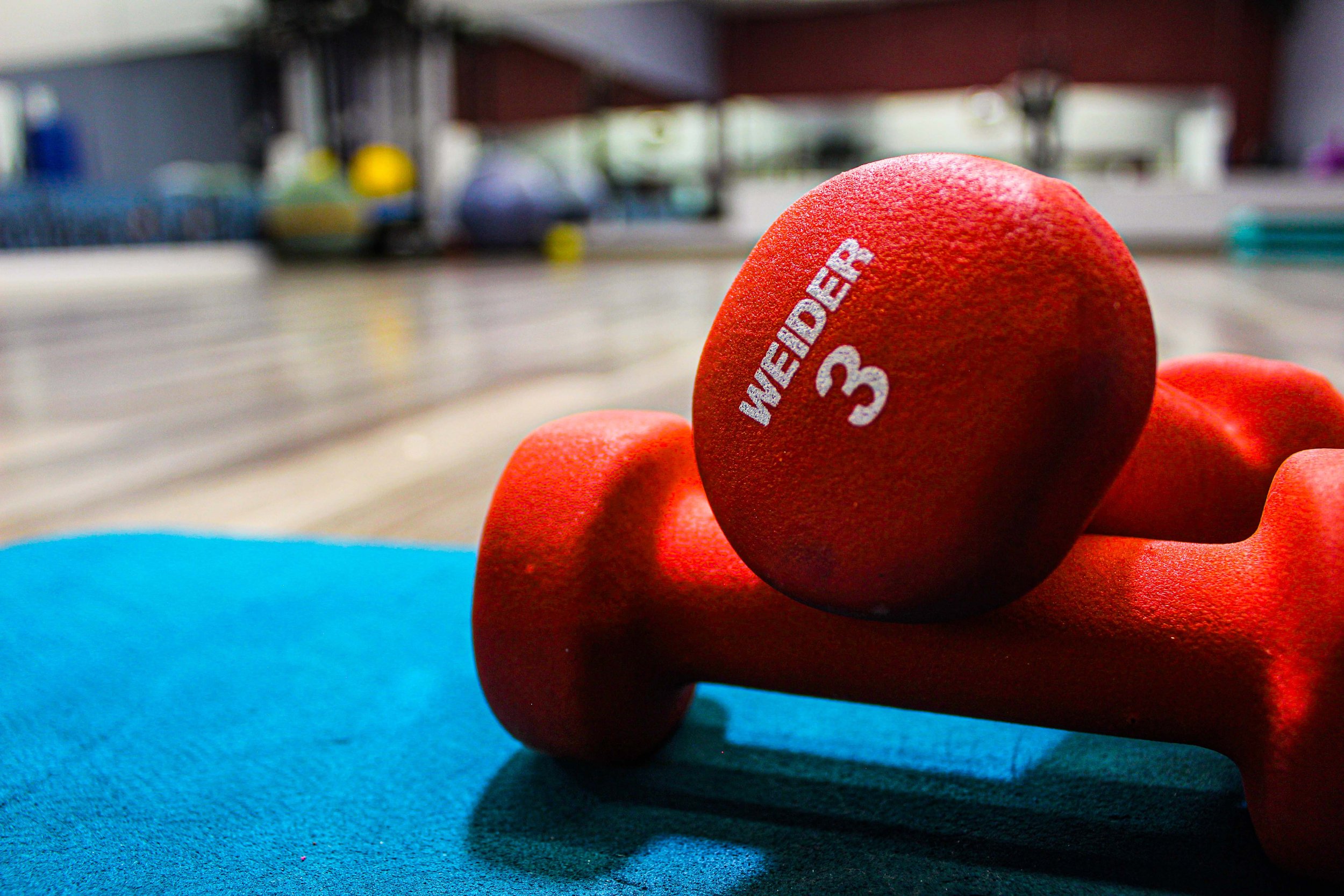 Close-up of a pair of red 3-pound Weider dumbbells resting on a blue workout mat in a brightly lit gym setting, with blurred gym equipment in the background.