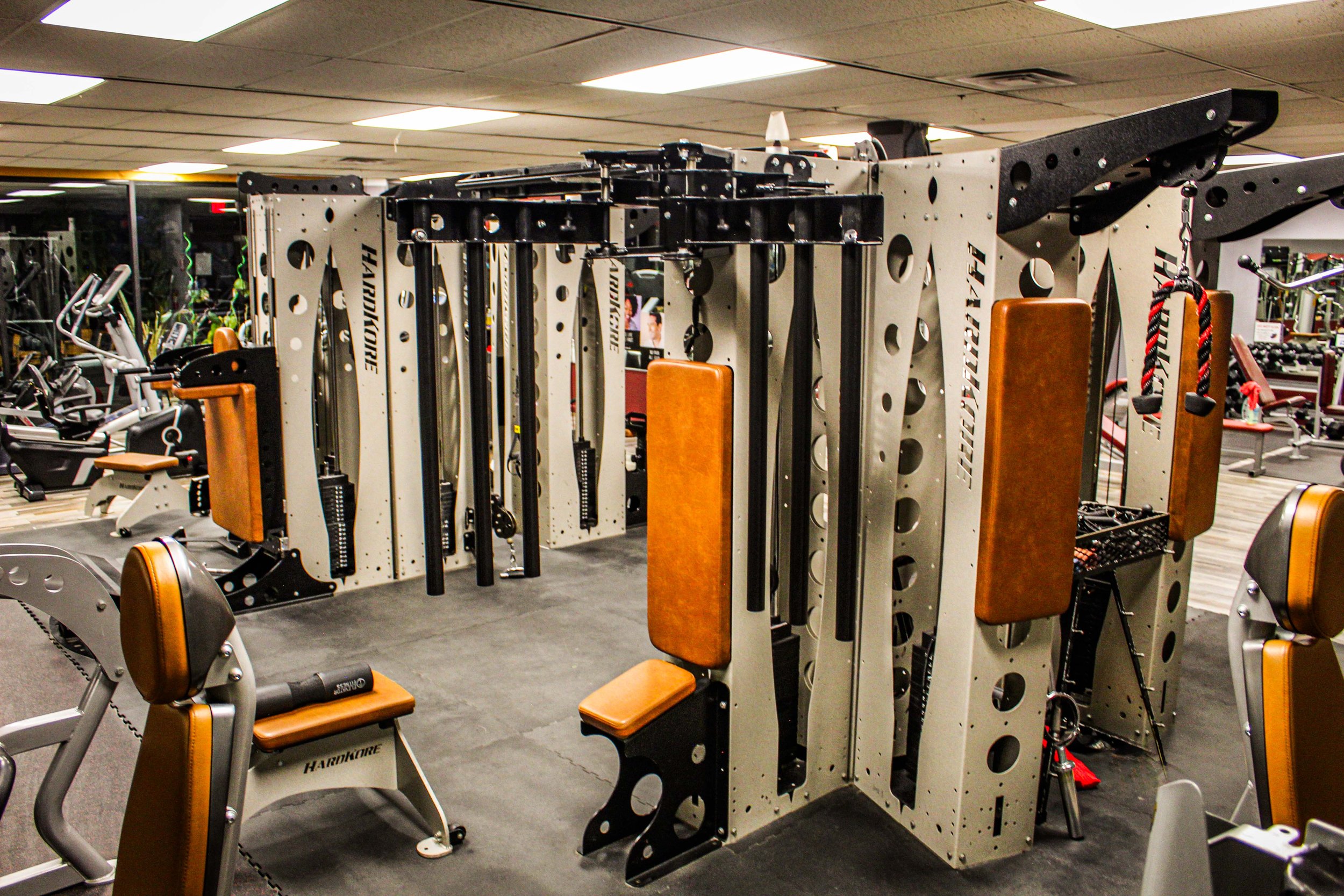 Multi-functional jungle gym equipment with brown padded seating and cable attachments, set in a gym with additional cardio and strength training machines in the background