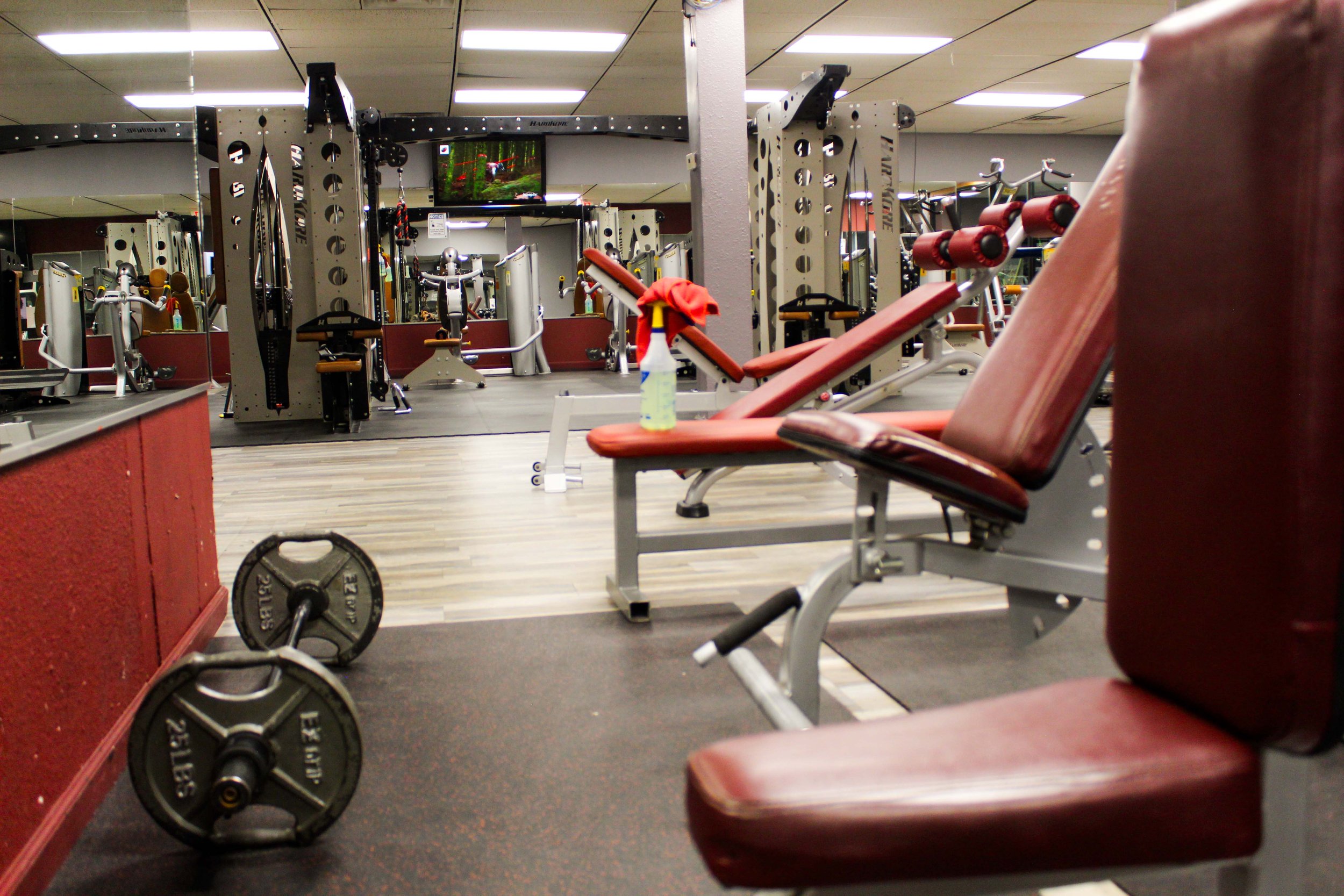 Workout benches in a gym with padded red seats, a weight bar on the floor, and a spray bottle for cleaning nearby. In the background, a mirrored wall reflects additional gym equipment, including weight machines and cable systems.