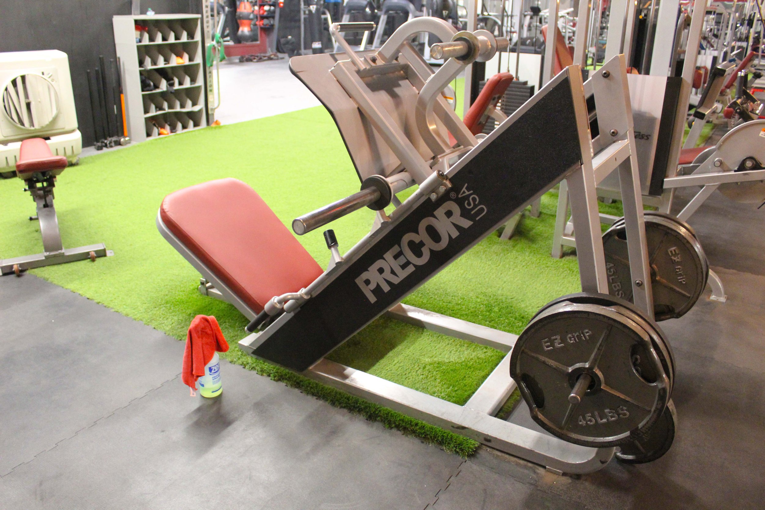 Precor leg press machine with 45-pound weight plates loaded, positioned on turf flooring in a gym with a red seat and nearby cleaning supplies.