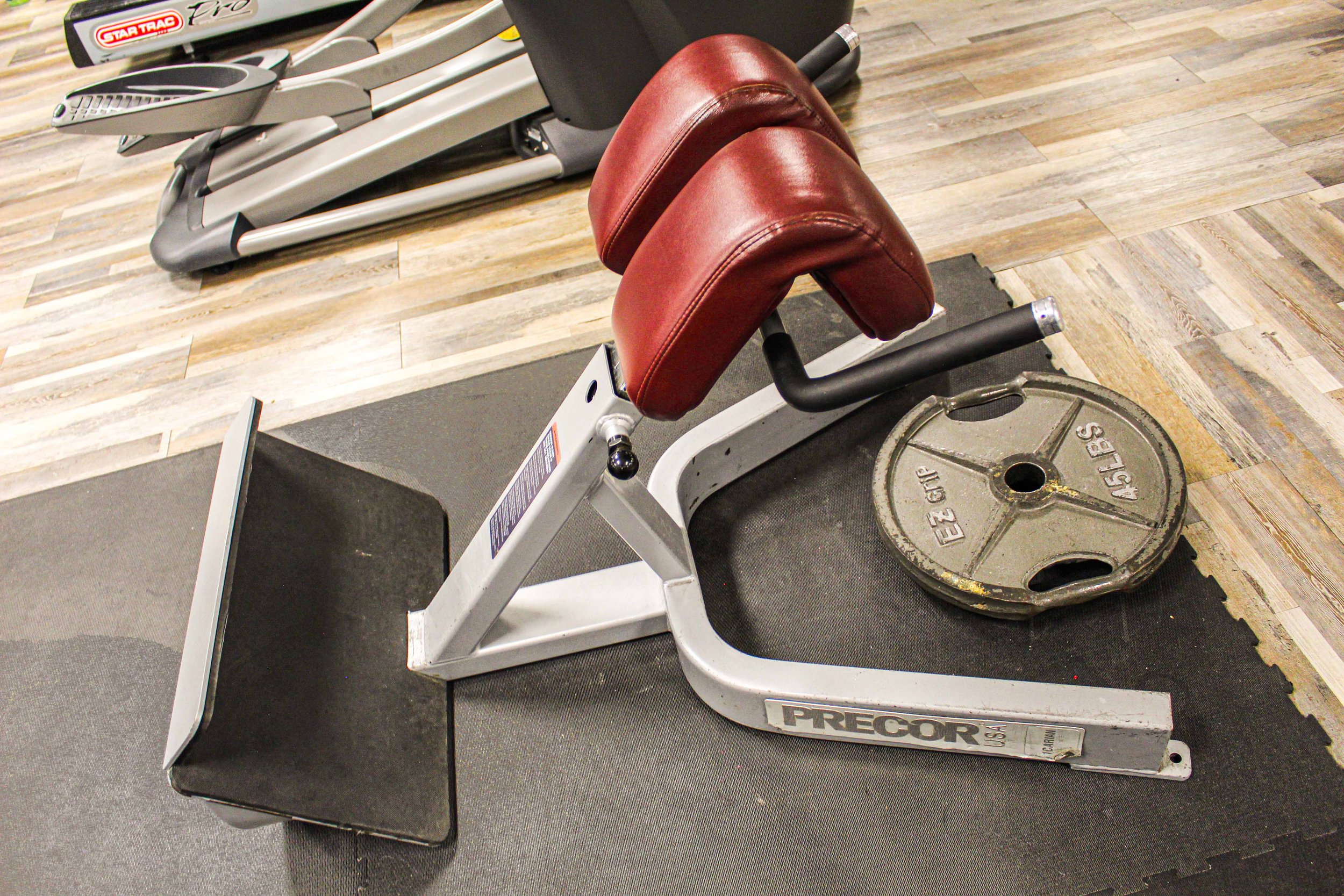 Back extension bench with red padded supports and a 45-pound weight plate placed nearby on a gym floor with wooden-style flooring