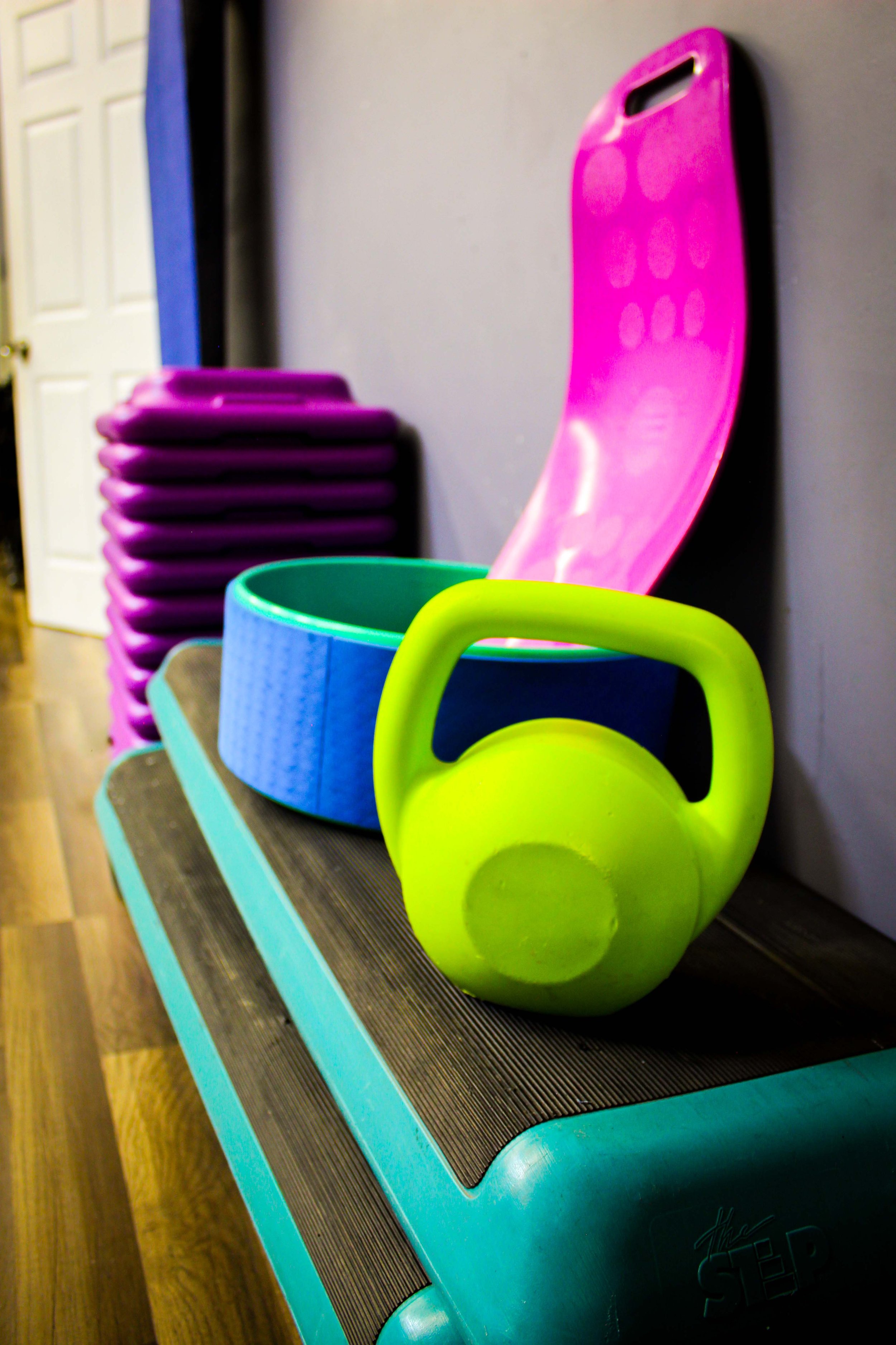 Brightly colored exercise equipment, including a green kettlebell, a blue foam wheel, a purple step riser, and a pink workout seat, placed on a wooden floor against a light purple wall.