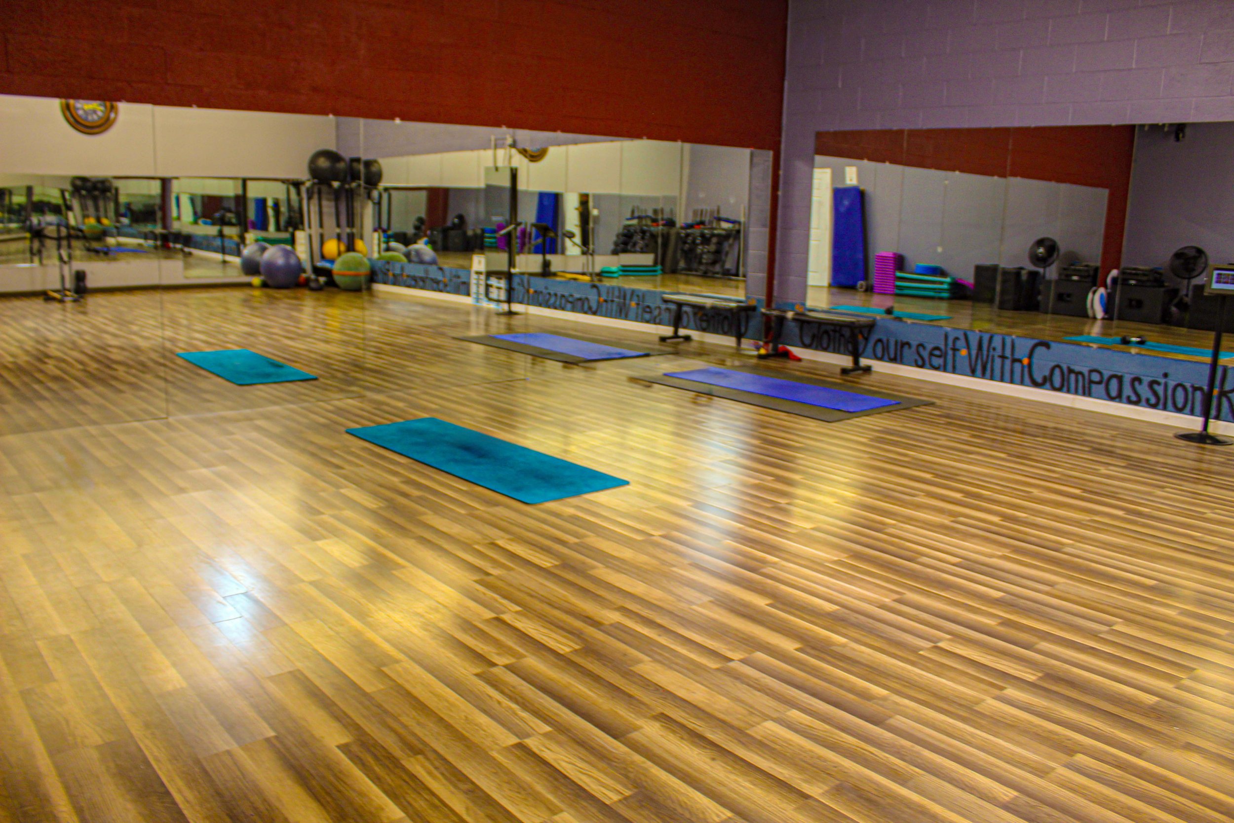 Spacious exercise room with wooden floors, yoga mats, mirrored walls, and fitness equipment like exercise balls and step platforms neatly arranged in the background.