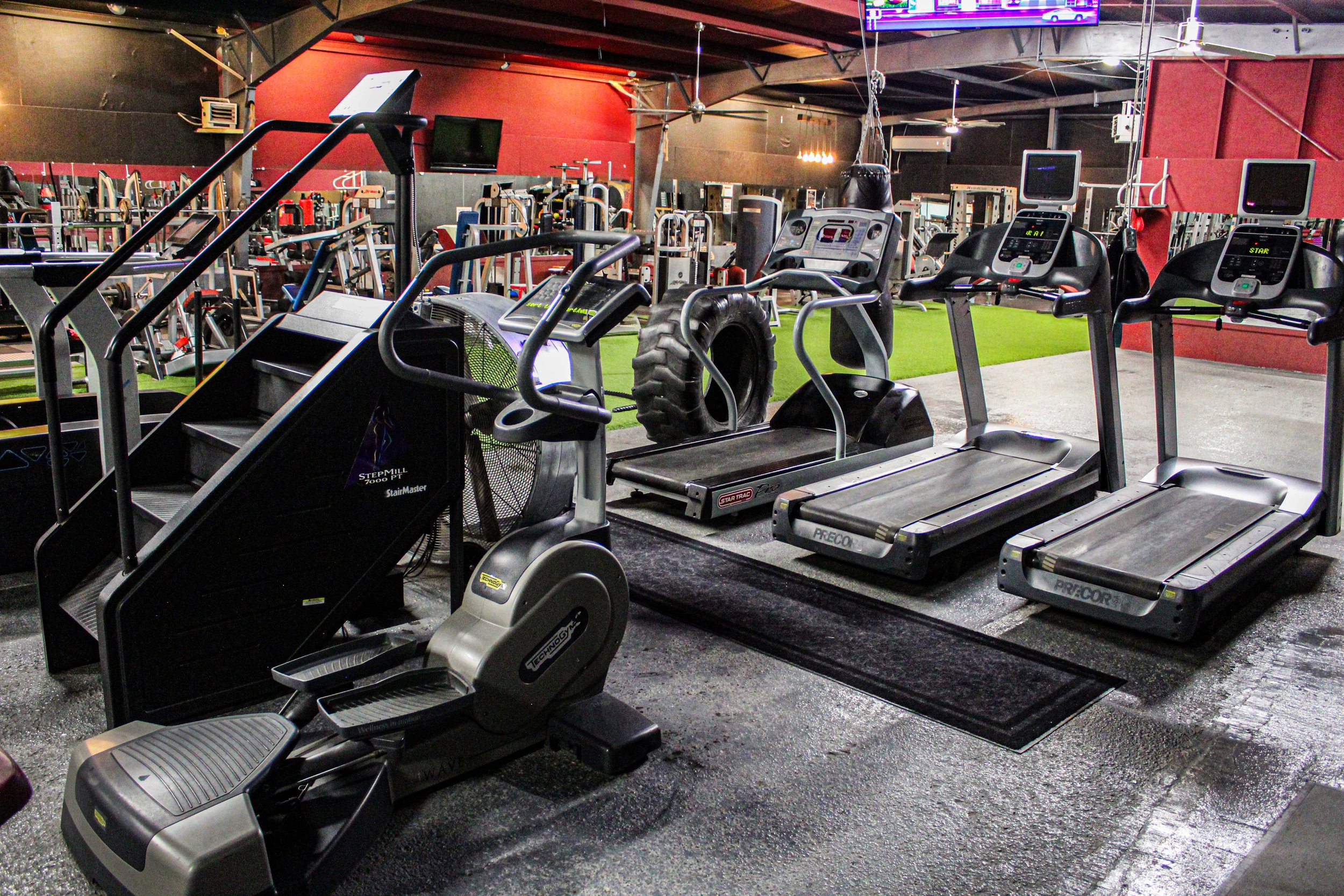 Cardio section in a gym featuring a StairMaster StepMill, elliptical trainer, and multiple treadmills, with gym equipment and a green turf workout area visible in the background.