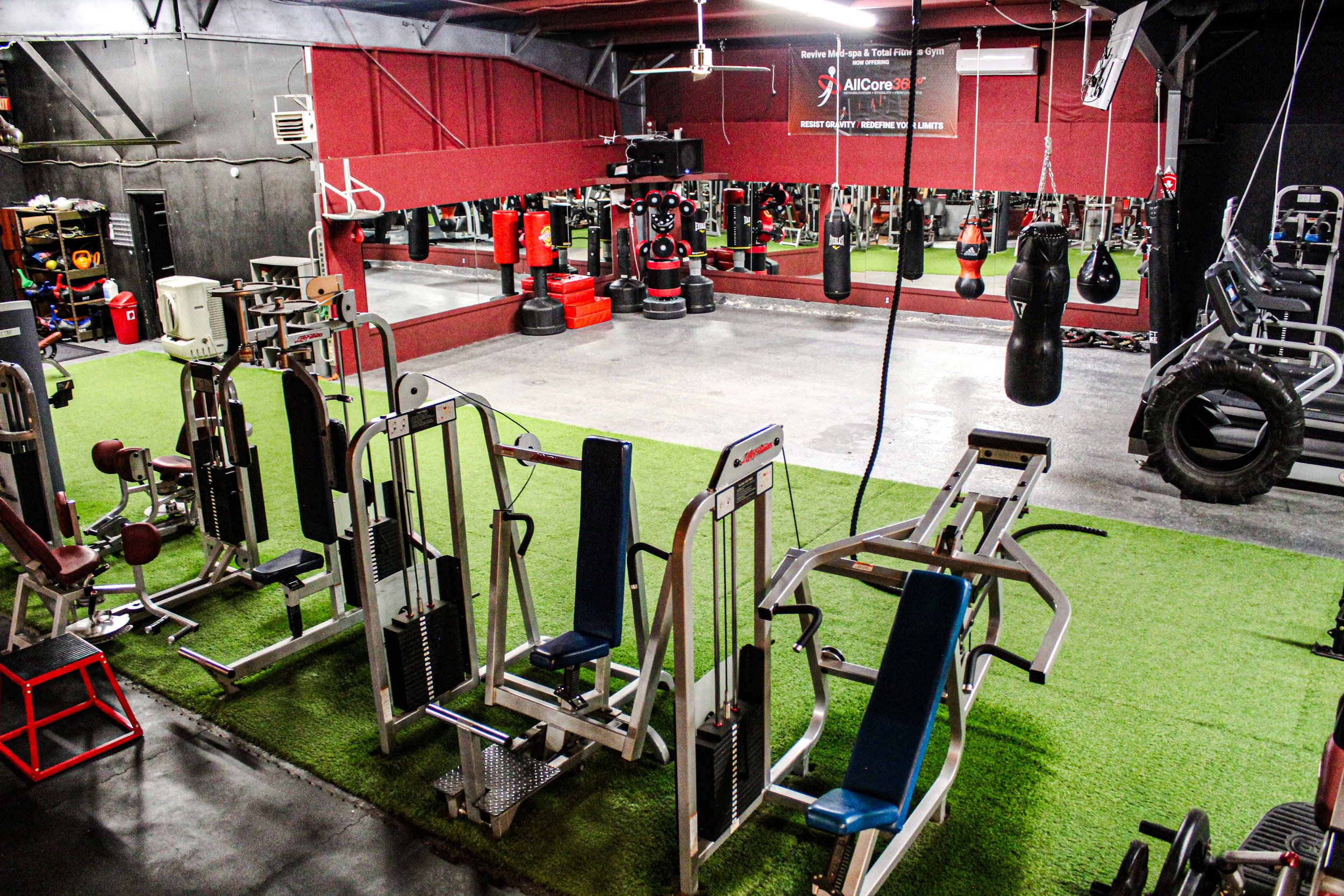 Overhead view of a gym featuring a boxing area with heavy bags, specialty striking equipment, and strength training machines on turf flooring, alongside a large tire and sledgehammer for functional workouts.
